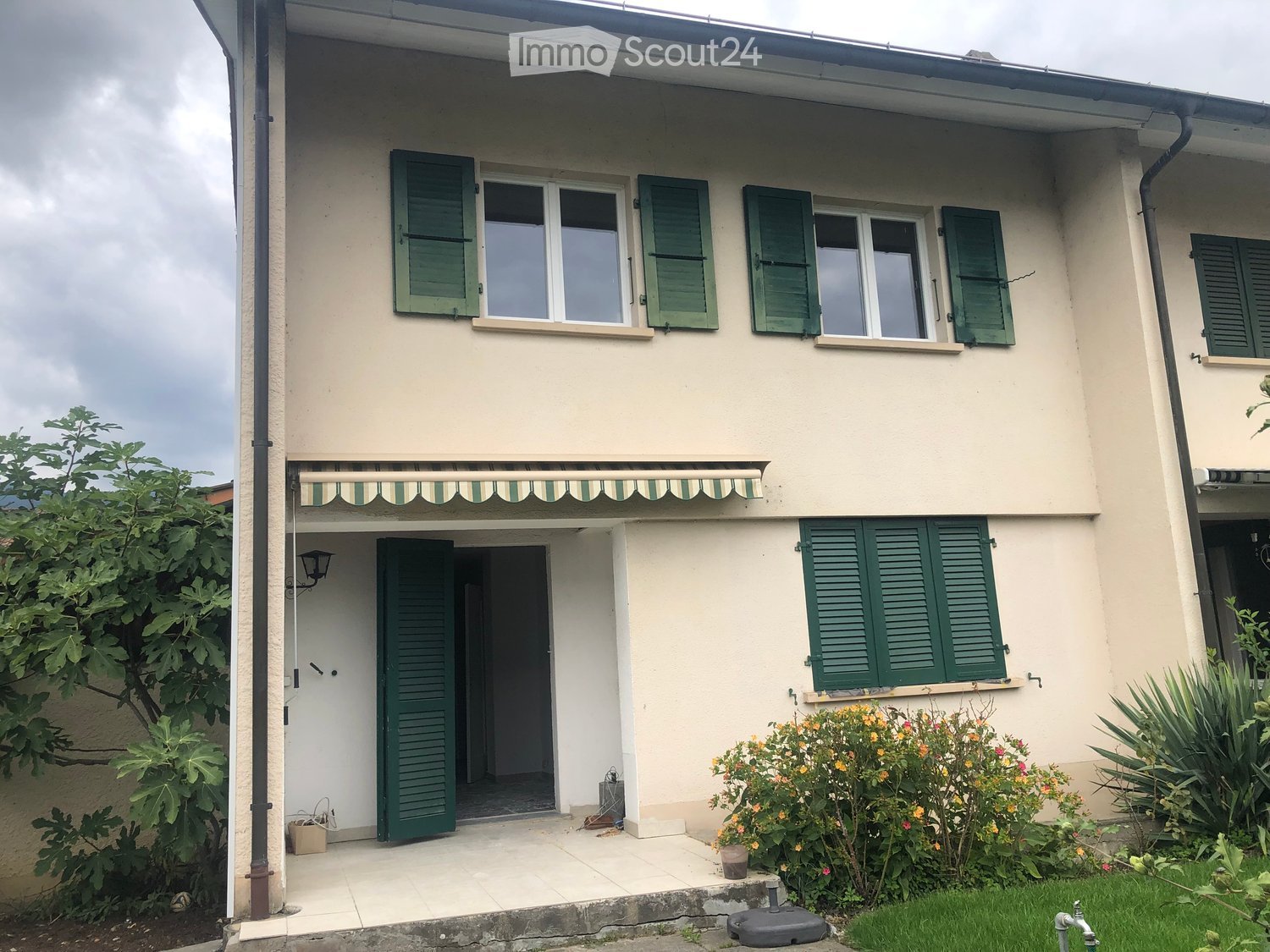 Two-story house with beige walls, green shutters, and a striped awning over the entrance. The front yard has potted plants and a small garden area.