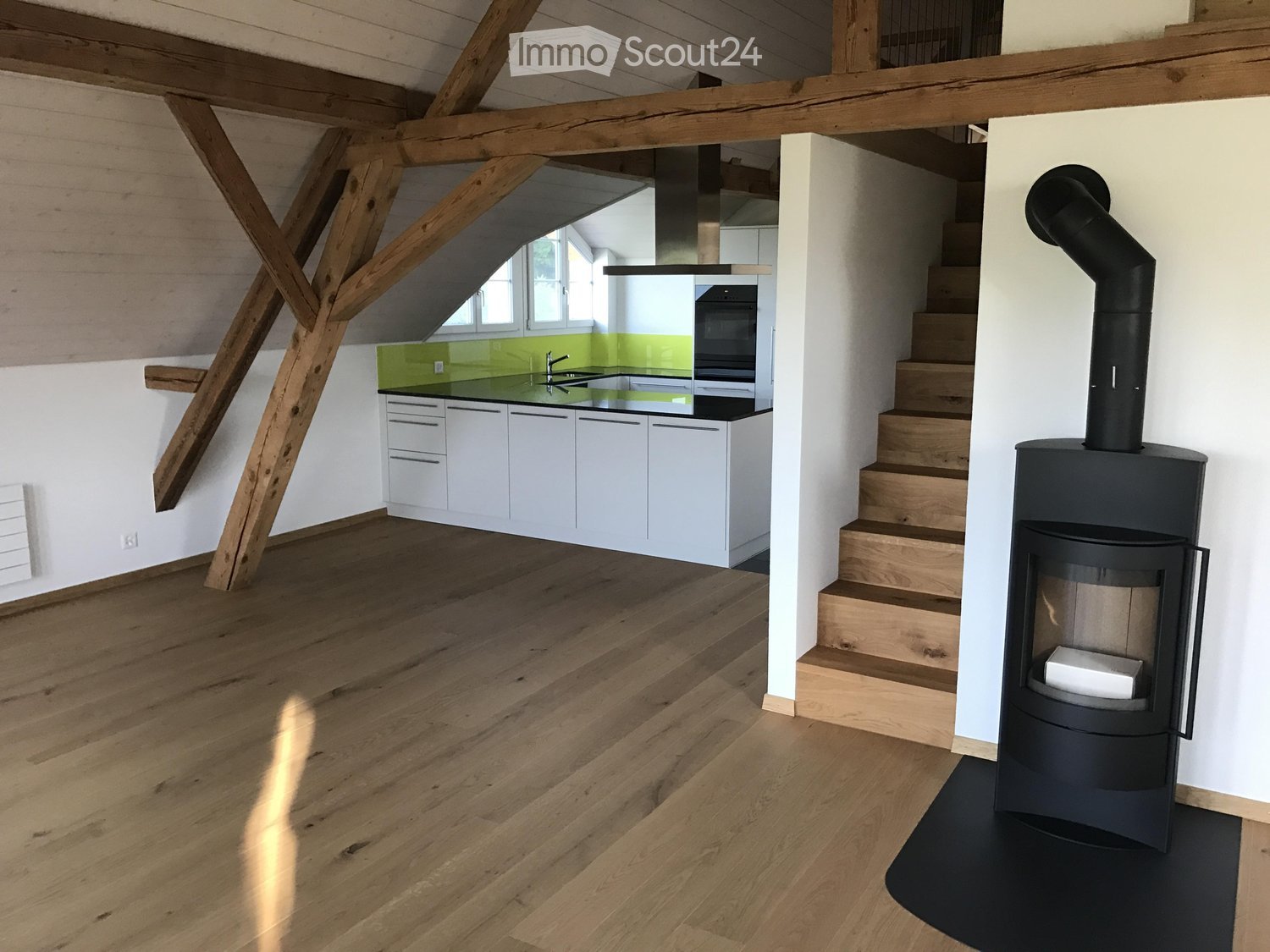 Open-plan space with exposed wooden beams, a modern kitchen area with white cabinets and a green backsplash, a staircase leading to an upper level, and a wood-burning stove with a chimney.