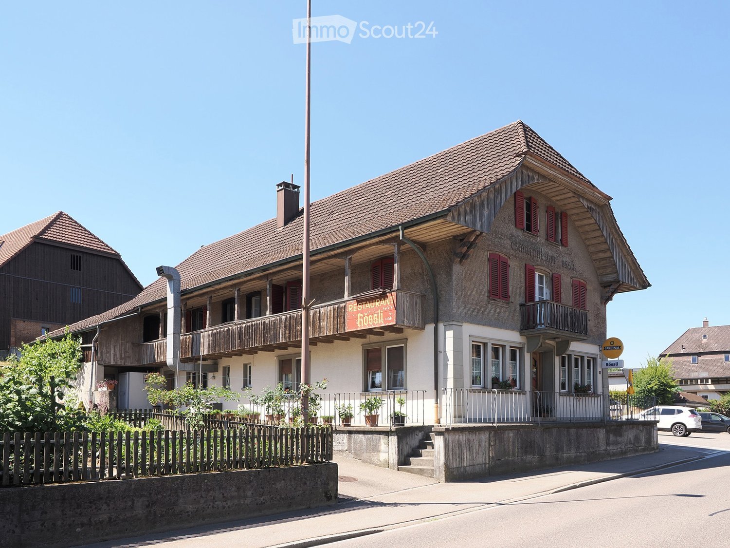 Two-story building with a restaurant sign "Restaurant Rossi" on the balcony, red shutters, and a wooden fence in front.