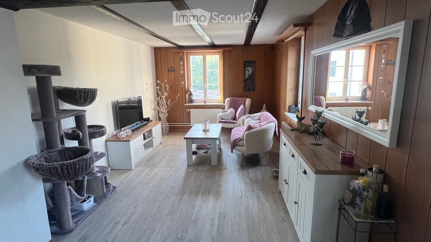 A cozy living room with wooden walls and flooring, featuring a cat tree, a cat litter, a small table with a candle, two chairs with pink cushions, a television on a cabinet, and a mirror on the wall.
