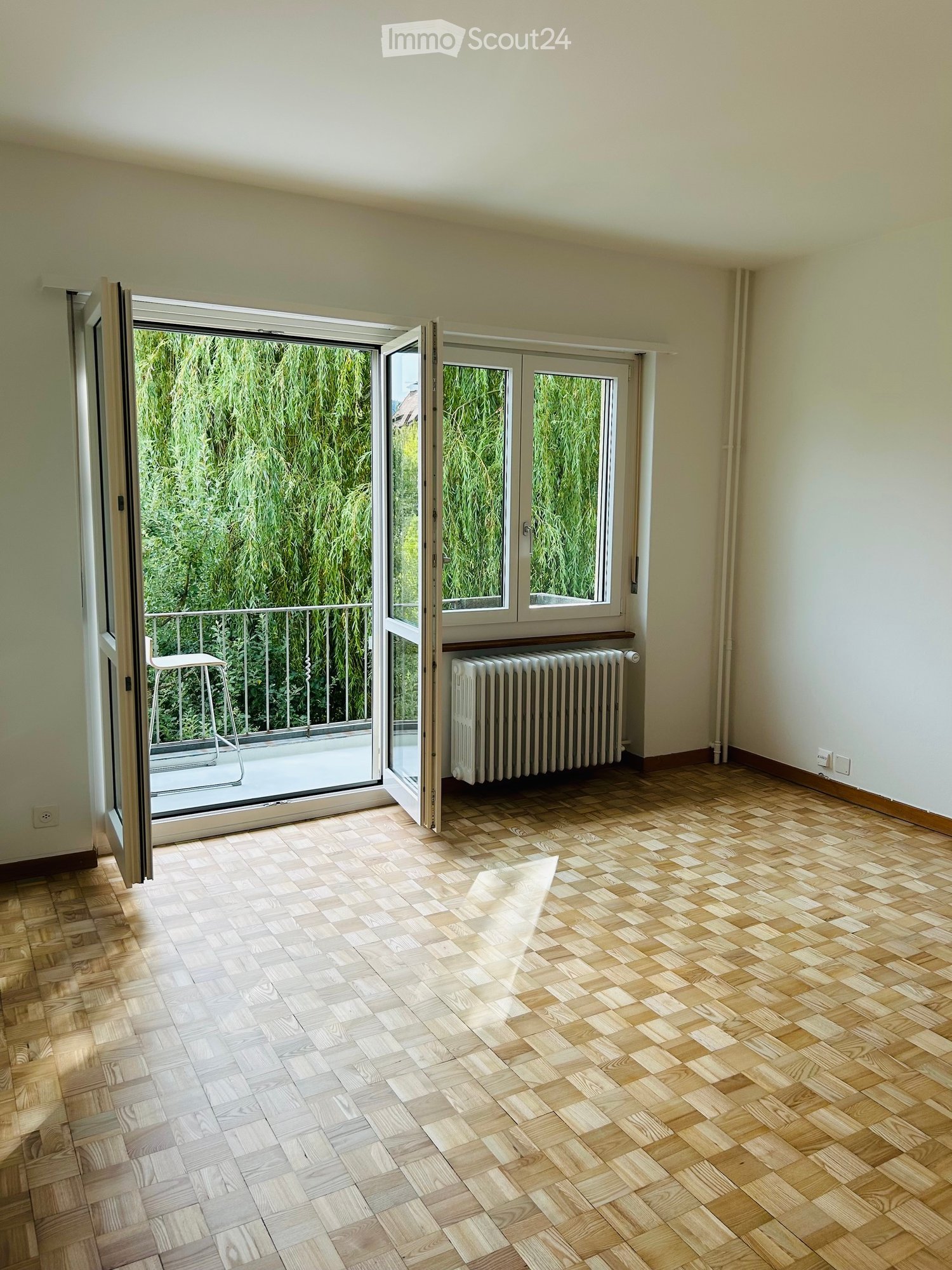 Empty room with wooden parquet flooring, white walls, large sliding glass doors leading to a balcony with a view of greenery, and a radiator under the window.
