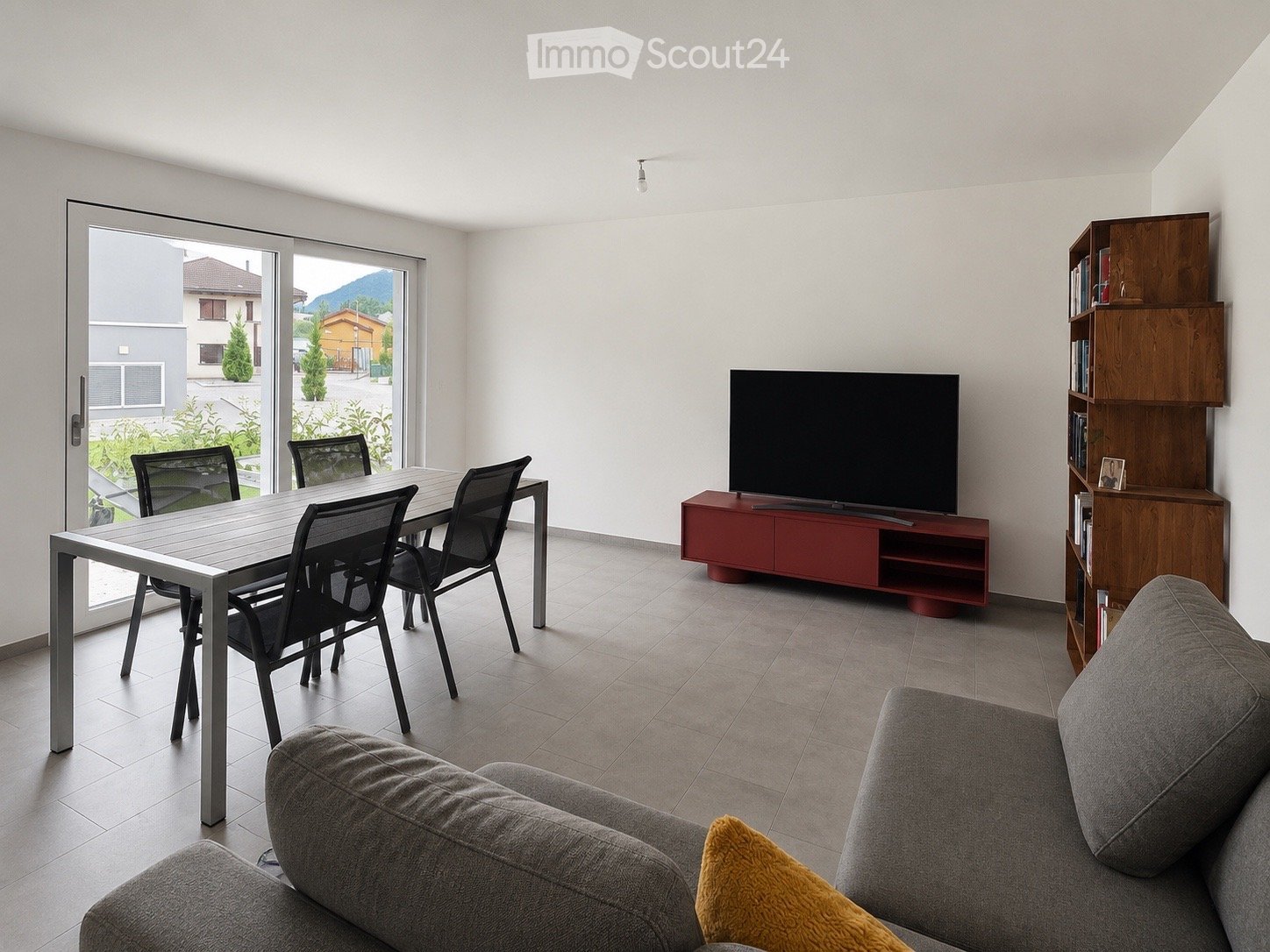 Modern living room with a grey sofa, a wooden shelf, a flat-screen TV on a red stand, a dining table with four chairs, and a large window with a view of the outdoor area.