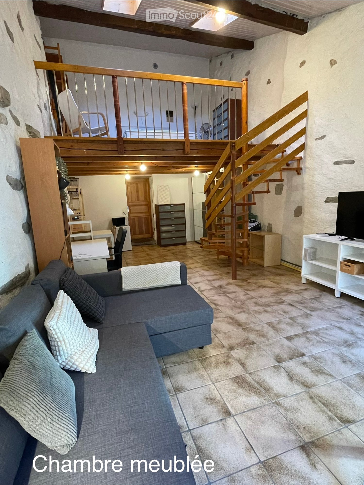 A furnished living space with a grey sectional sofa, a wooden staircase leading to an upper level, a TV on a white stand, a small table with chairs, and a fan in the background.