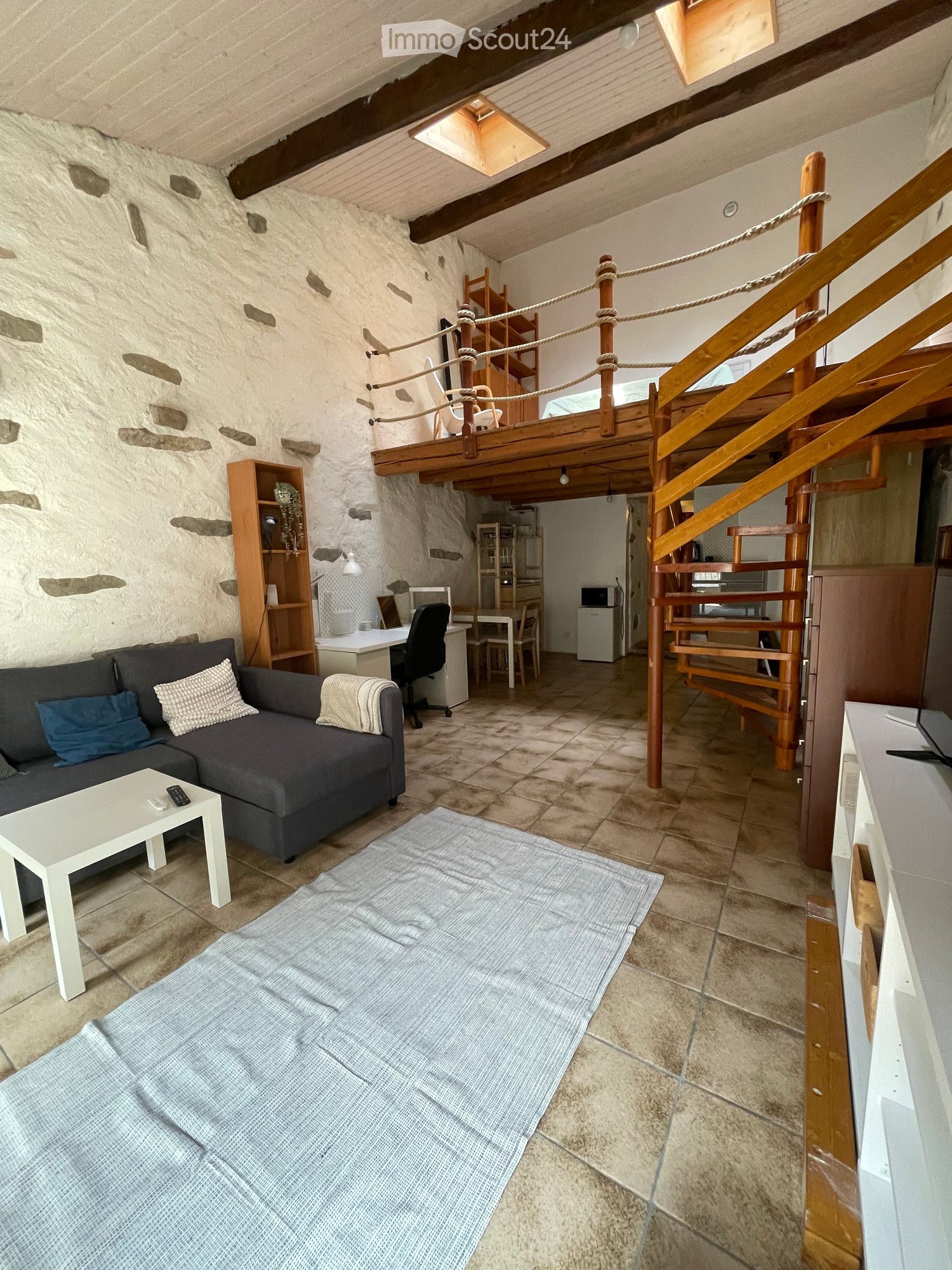 A cozy, rustic living space with a stone wall, a raised wooden bed platform, a sofa, coffee table, and a small workspace area with a desk and chair.