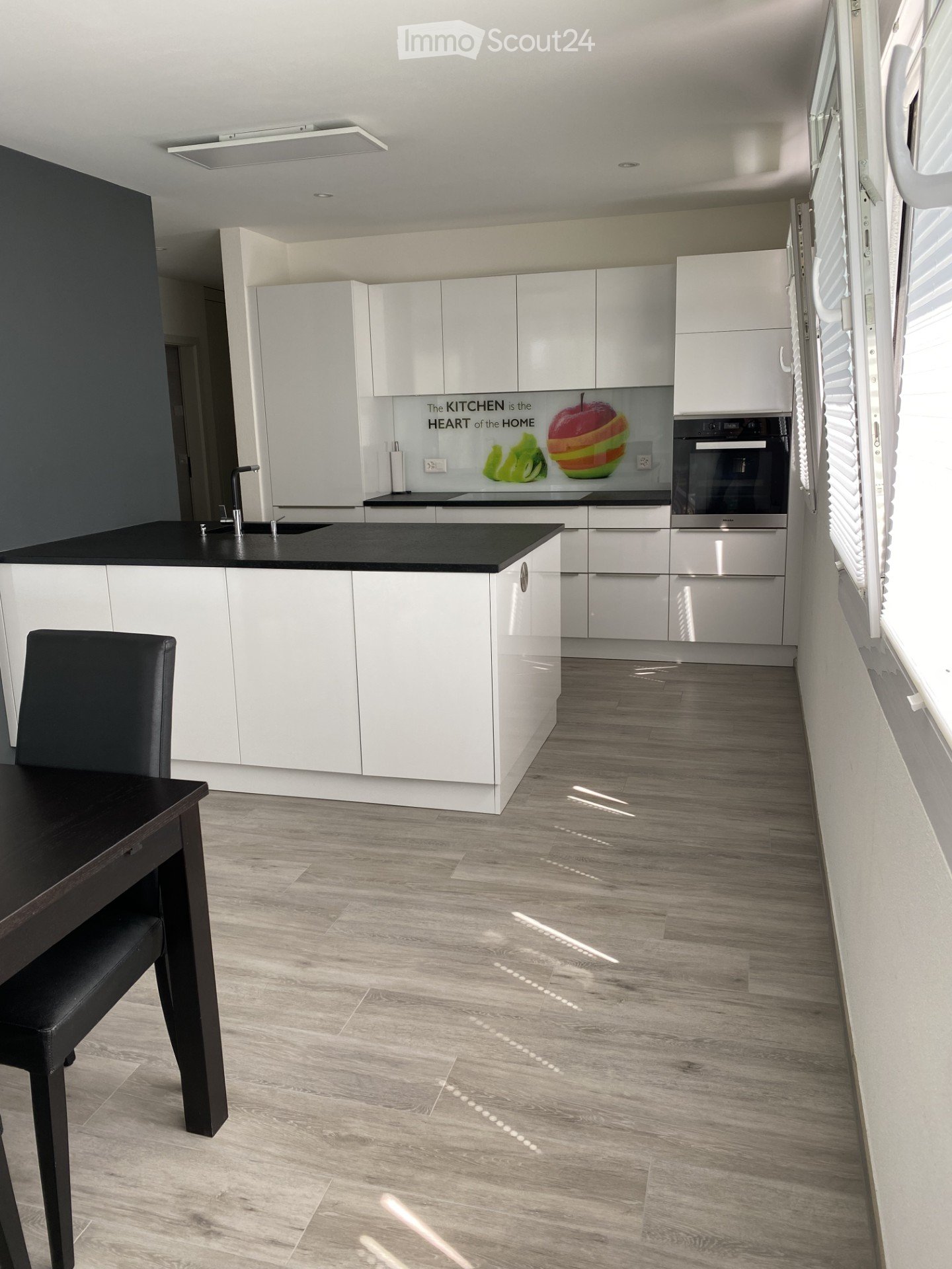 Modern kitchen with white cabinets, black countertop, and a sleek design. Features a built-in oven, sink with faucet, and a small dining area with a black table and chair.