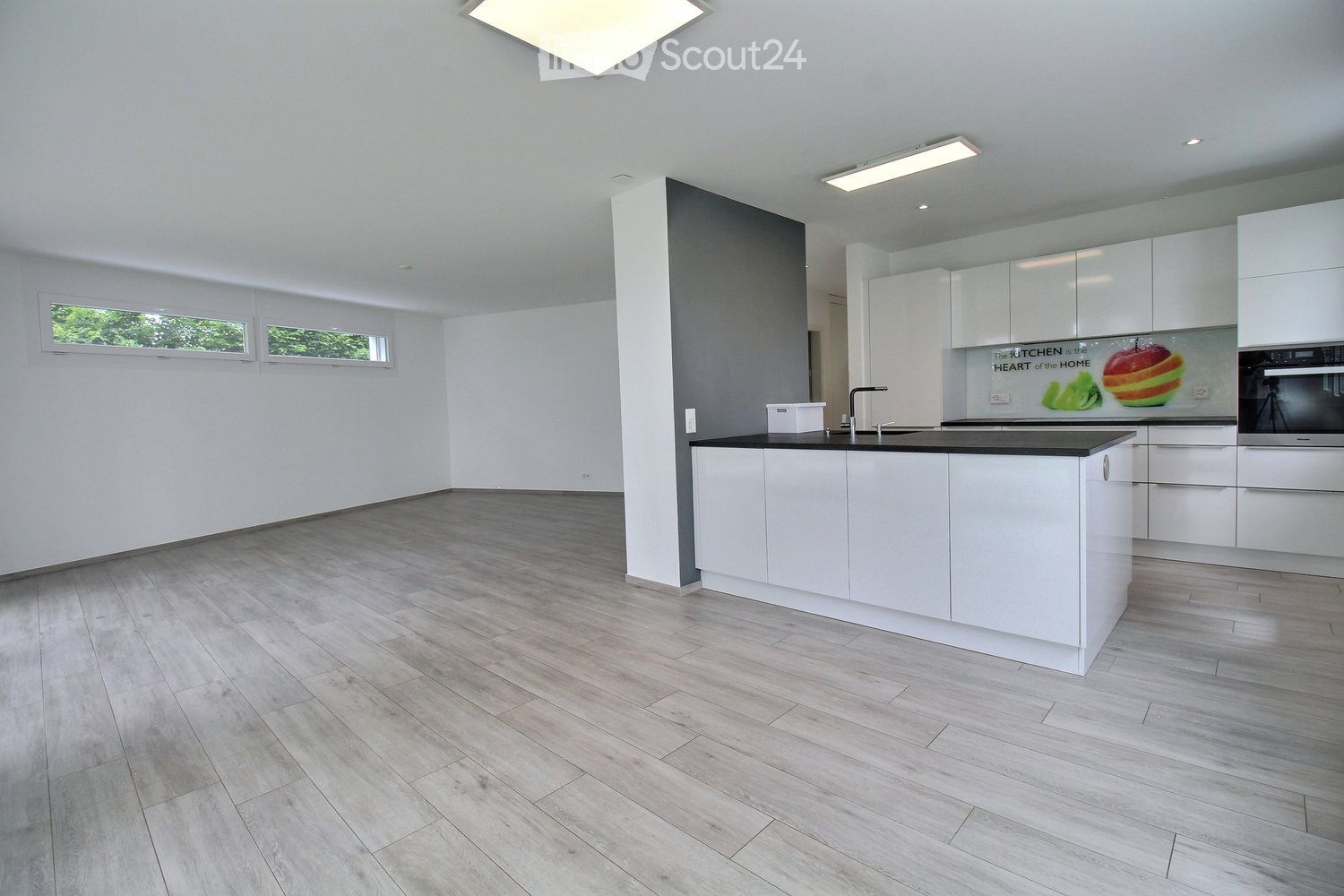 Modern, minimalist kitchen with white cabinets, a central island, and a sleek black countertop. The space features light wood flooring and large windows allowing natural light to fill the room.