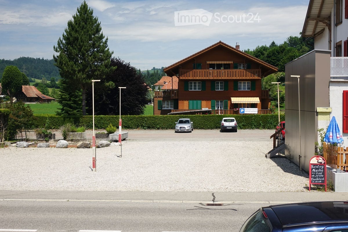 A large wooden house with green shutters and a balcony, surrounded by trees and a parking area with several cars.