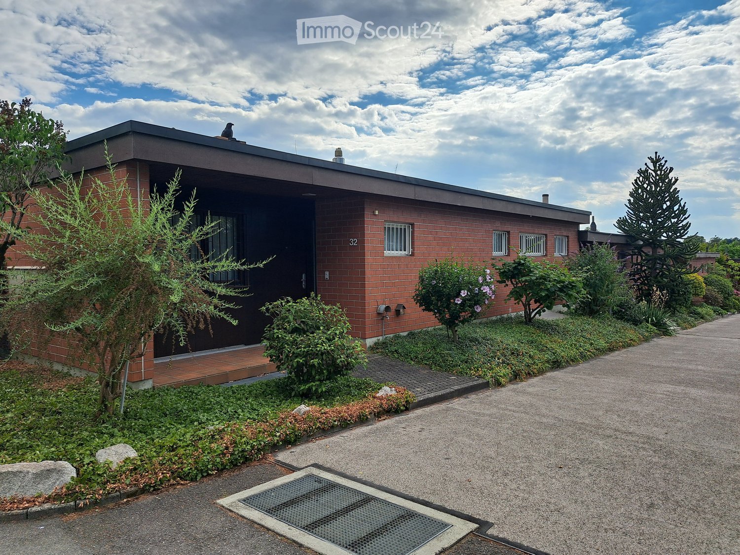 Single-story brick house with a flat roof, surrounded by landscaped garden with shrubs and trees. The entrance features a small porch with a tiled floor and a bird perched on the roof.