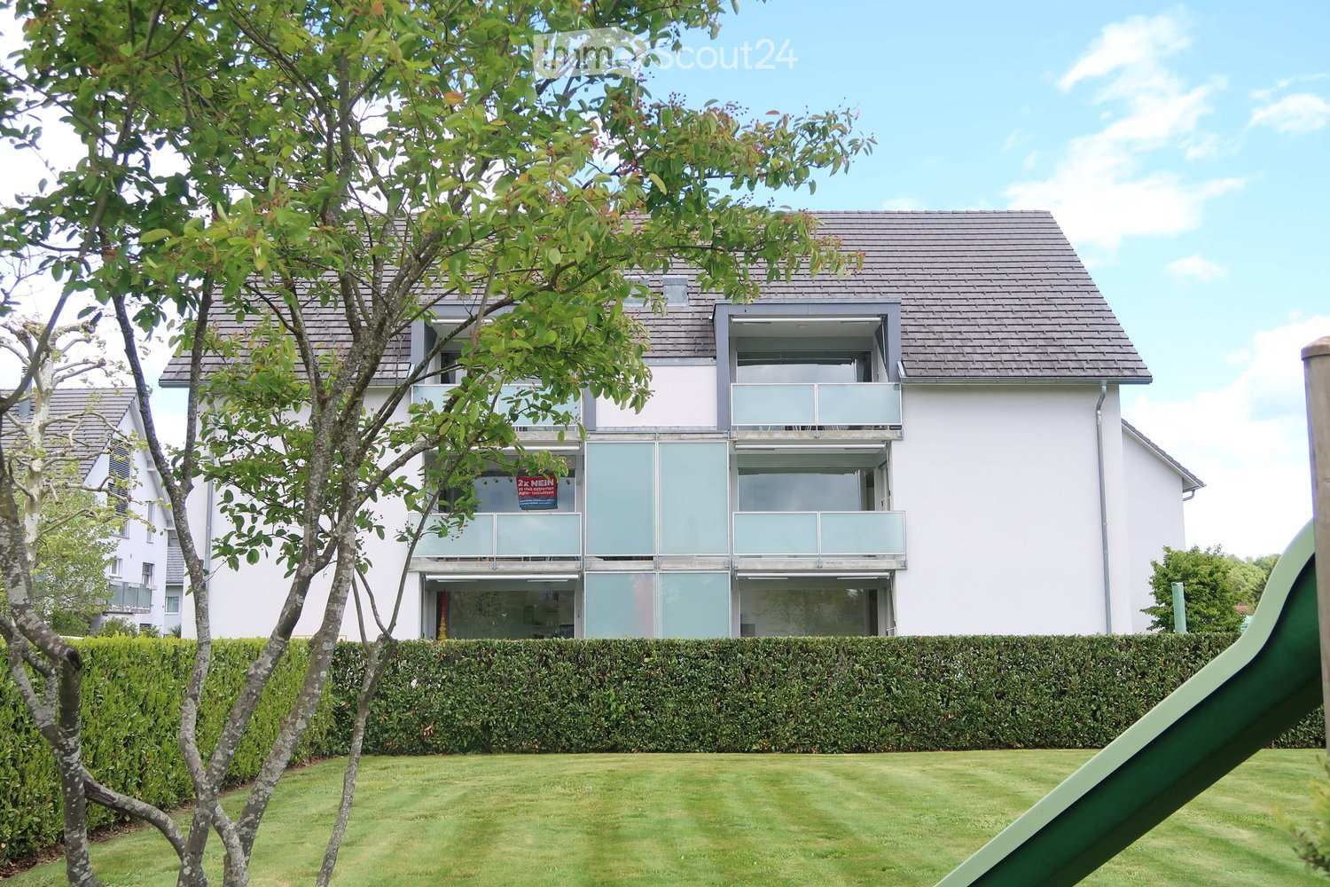 Modern two-story house with white exterior, large windows, and balconies. The house features a well-maintained lawn and a hedge in the front yard.