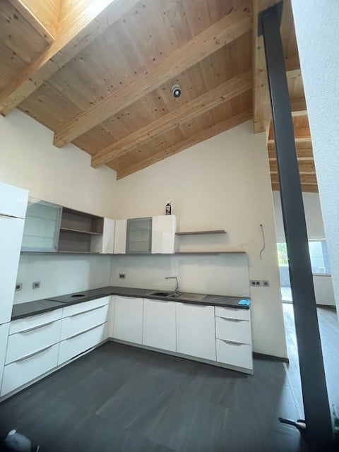 Modern kitchen with white cabinets, dark countertops, and a sleek design. Features a wooden slanted ceiling and a stainless steel sink.