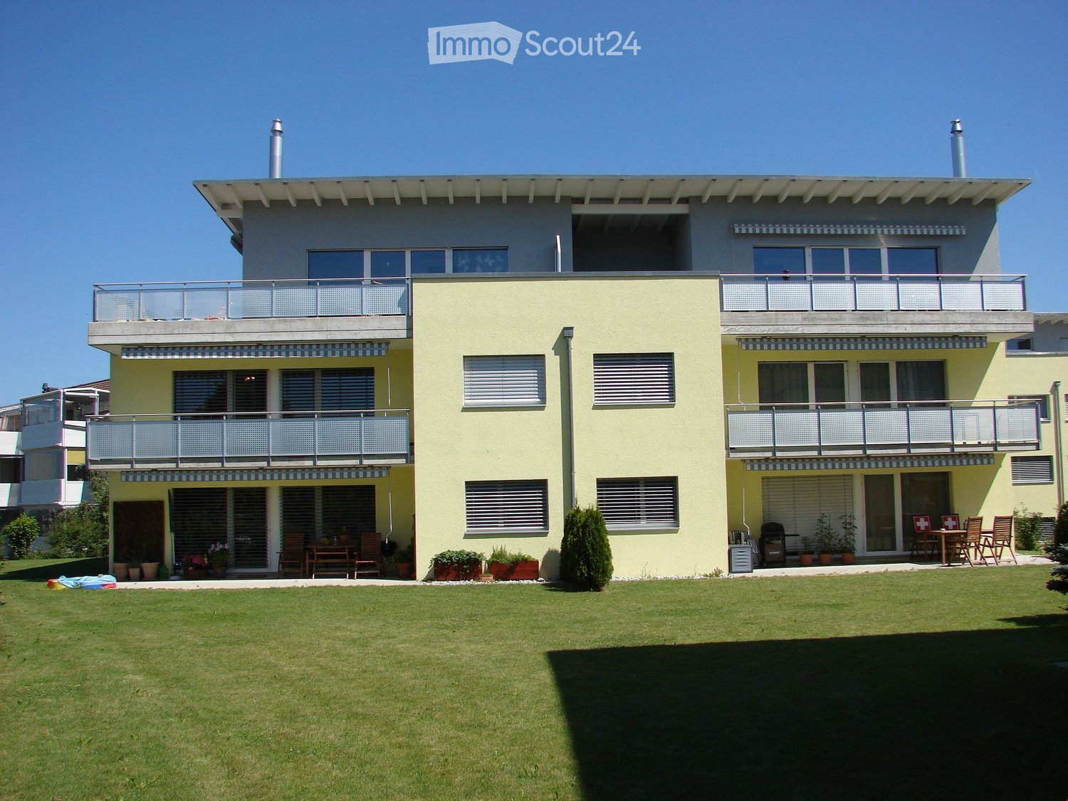 Two-story house with yellow exterior, multiple balconies, large front lawn, patio furniture, and potted plants.