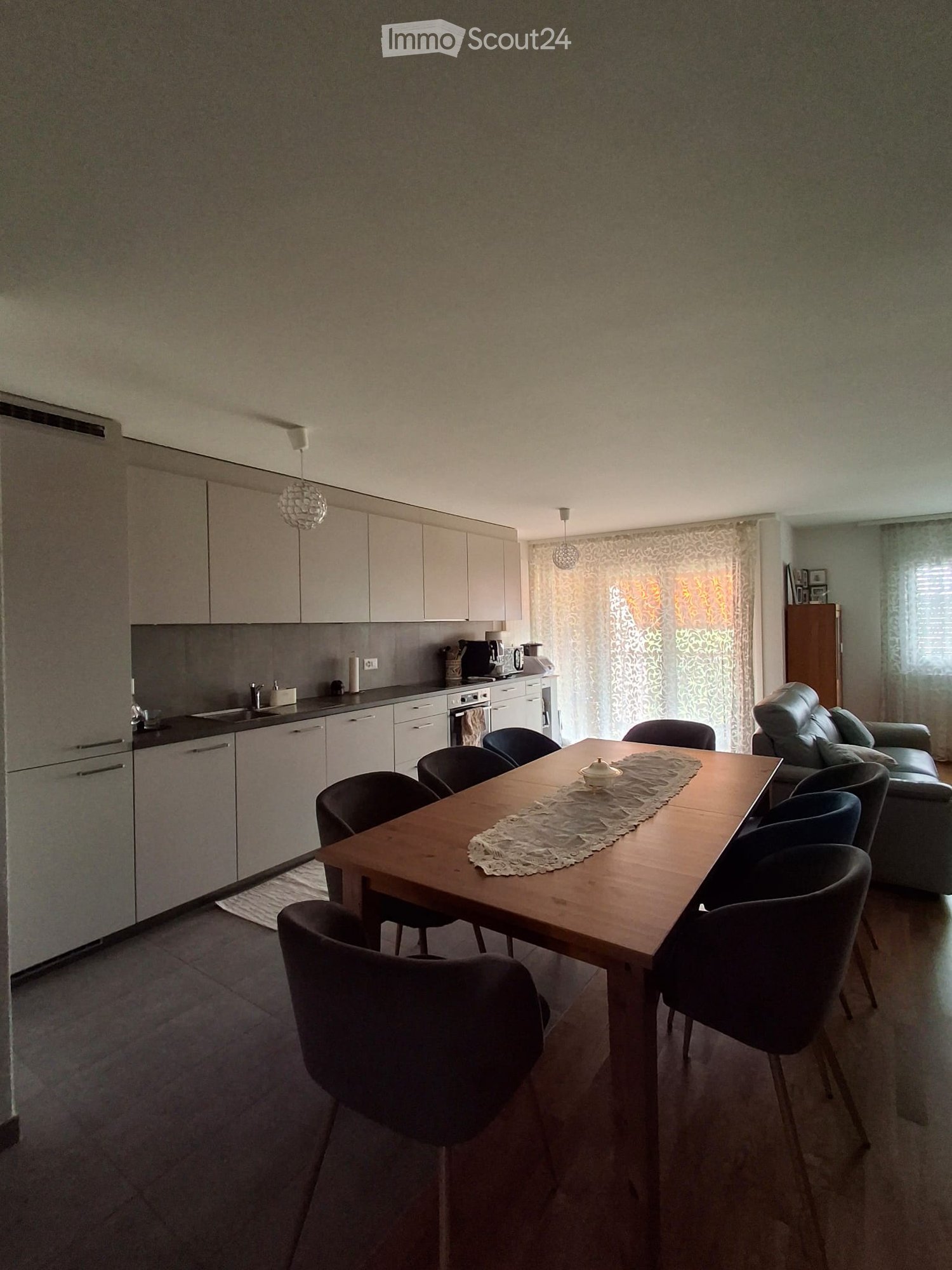 A modern, open-plan living and dining area with a wooden dining table, six chairs, a sofa, and a kitchen area with cabinets, a refrigerator, and an oven.