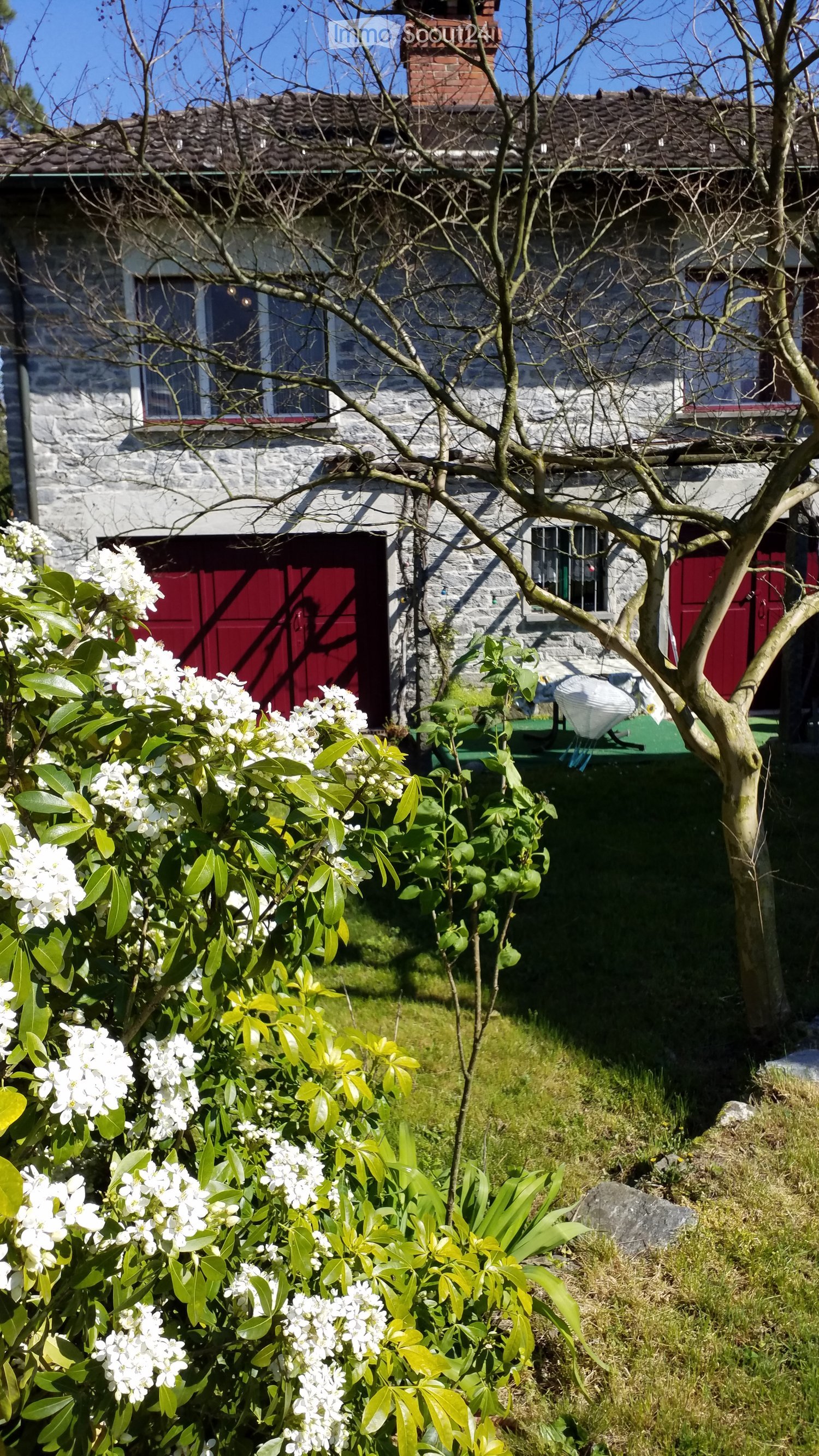 A stone house with red doors and windows, surrounded by a garden with flowering shrubs and a small tree.