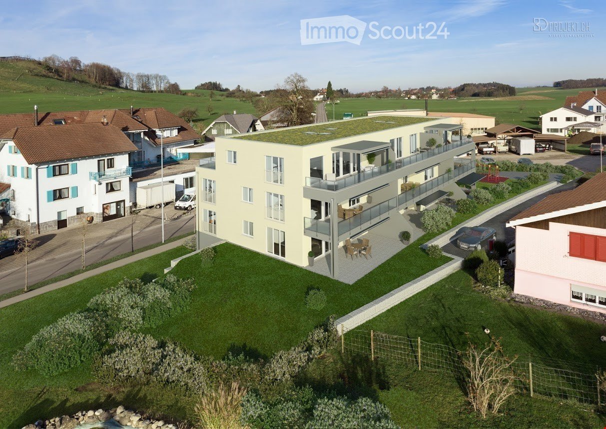 Modern multi-story building with a green roof, large windows, and a spacious balcony area. The surrounding area includes a well-maintained lawn, a parking lot, and neighboring houses.
