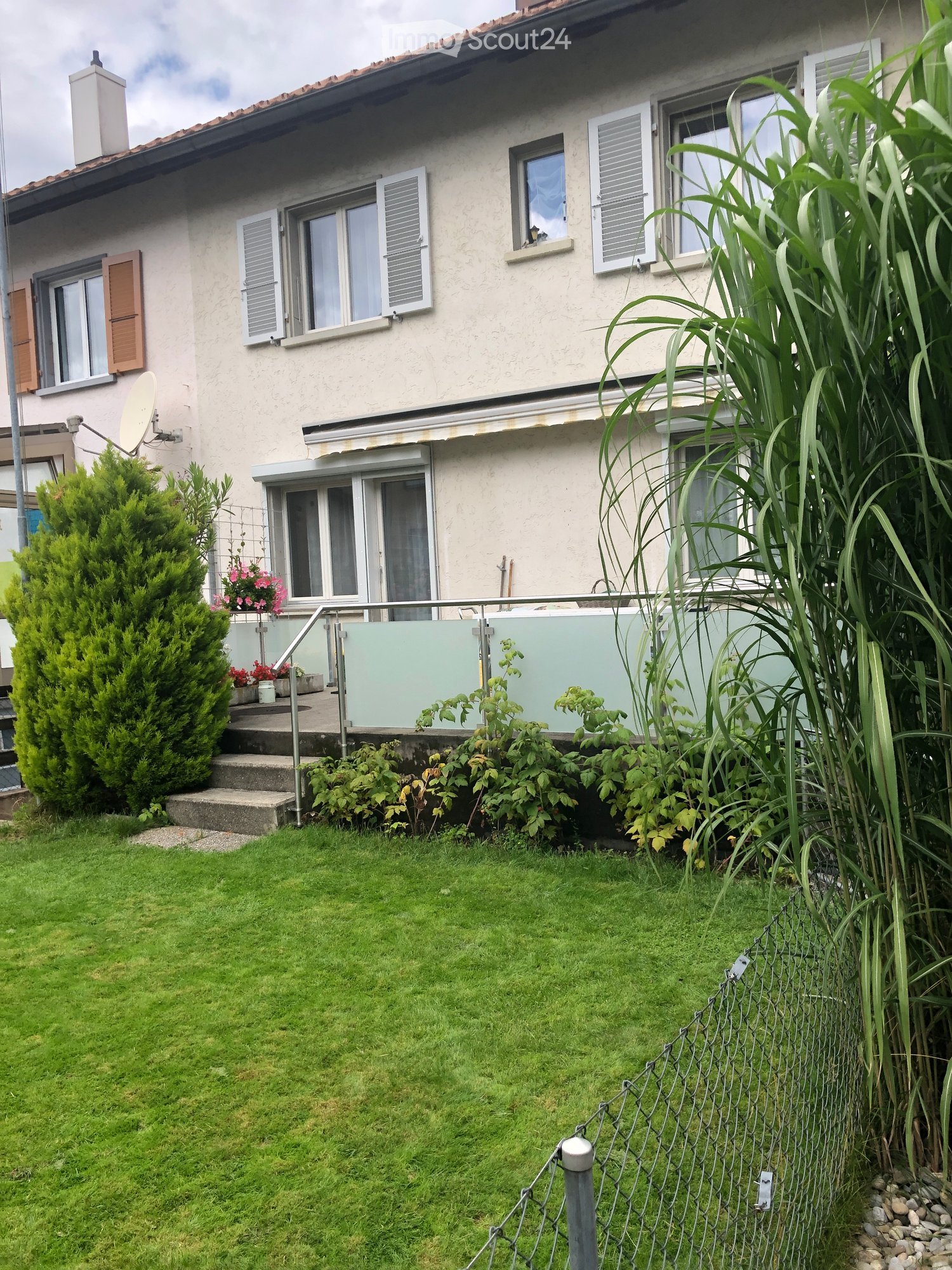 Two-story house with beige exterior, multiple windows with shutters, a chimney, and a satellite dish. The house has a well-maintained garden with green grass, shrubs, and a small staircase leading to a covered patio area.