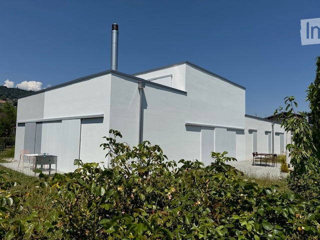 Modern, minimalist white building with a flat roof and chimney, surrounded by greenery and outdoor seating.