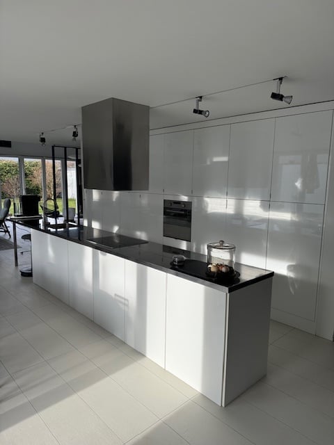 Modern kitchen with white cabinetry, black countertop, built-in oven, range hood, and a small dining area with a table and chairs.
