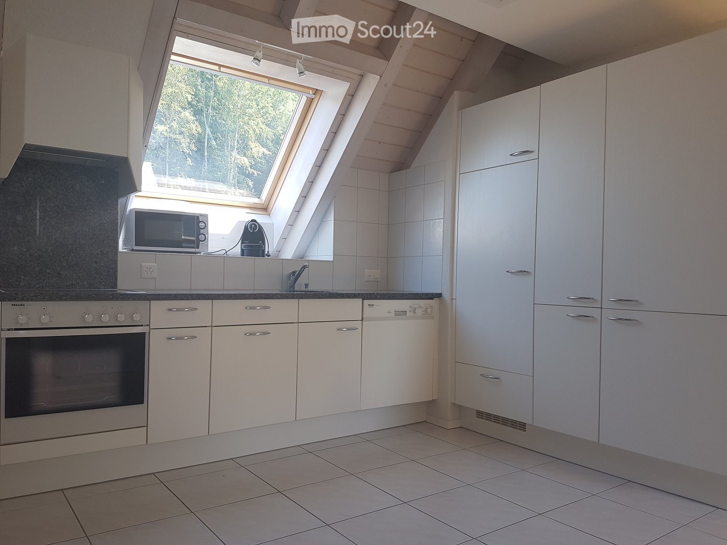Modern kitchen with white cabinets, tiled floor, and stainless steel appliances including an oven and microwave. Features a skylight window above the sink.