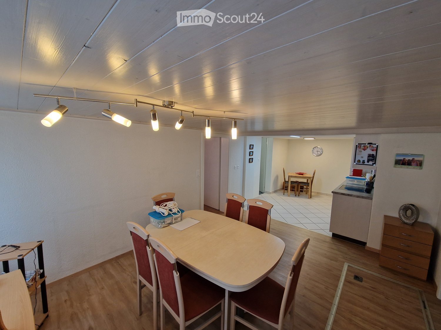 Bright dining area with a round wooden table, six chairs, a small cabinet, and a clock on the wall. The room has wooden flooring and recessed lighting.
