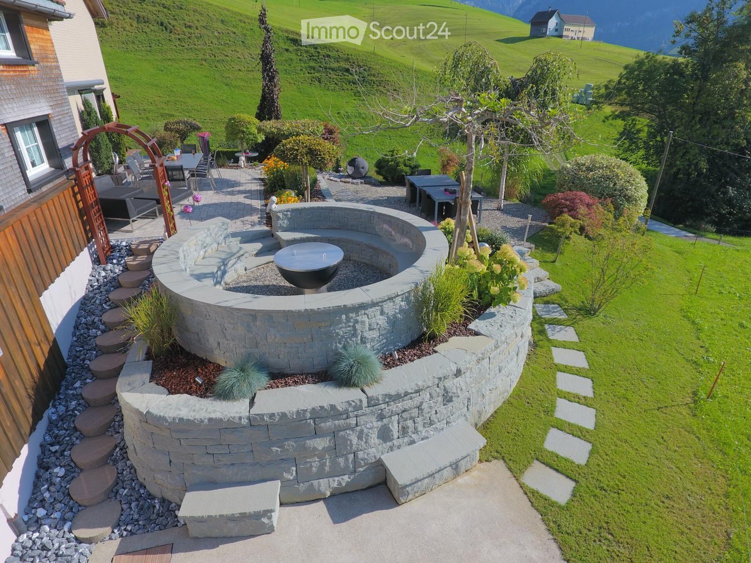 A well-maintained garden with a stone fire pit, surrounding seating area, and lush greenery, featuring a view of a green hillside and distant mountains.
