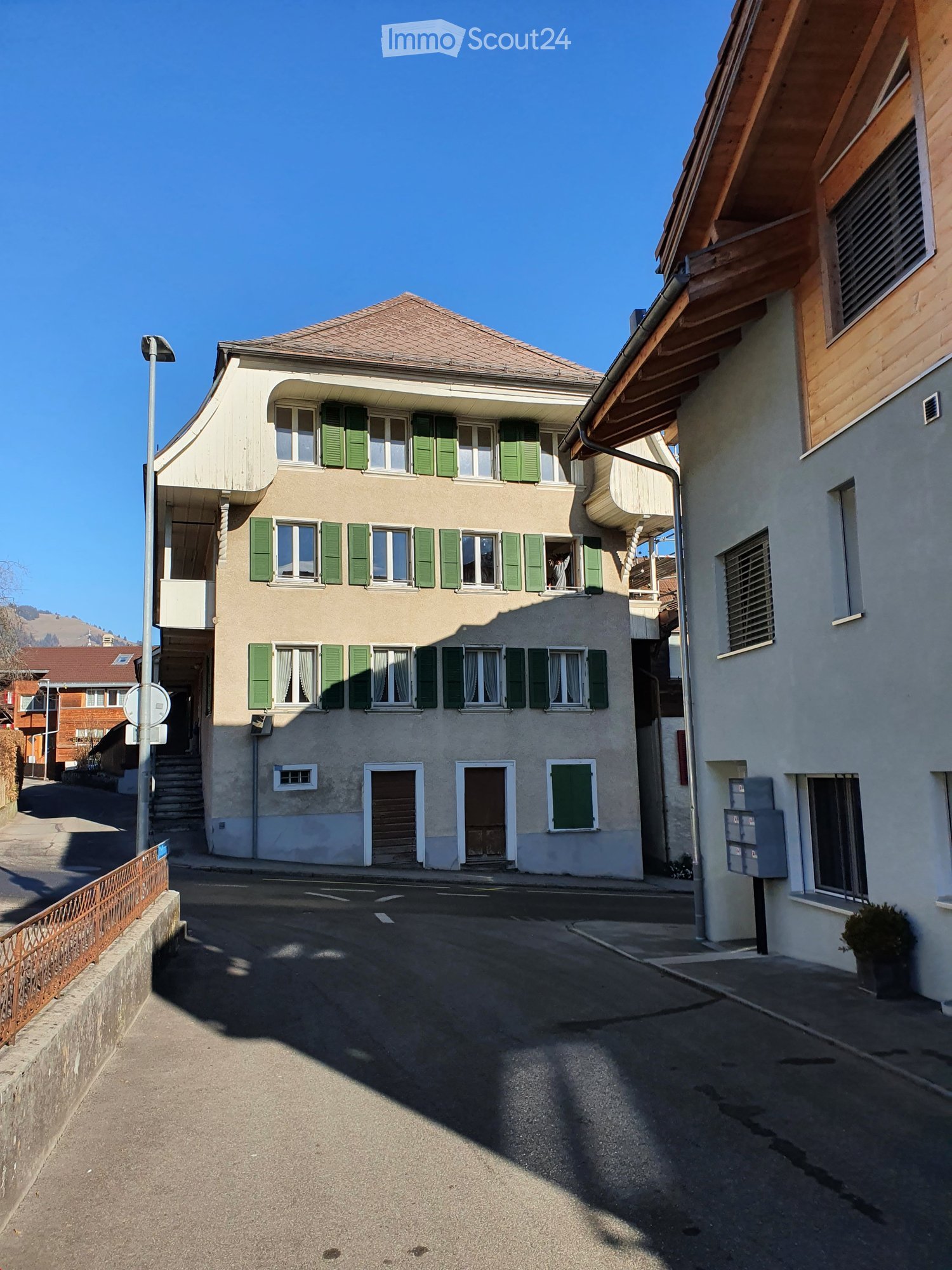 A three-story residential building with beige walls, green shutters, and multiple windows. The building features a small balcony on the second floor and a staircase leading to the entrance.