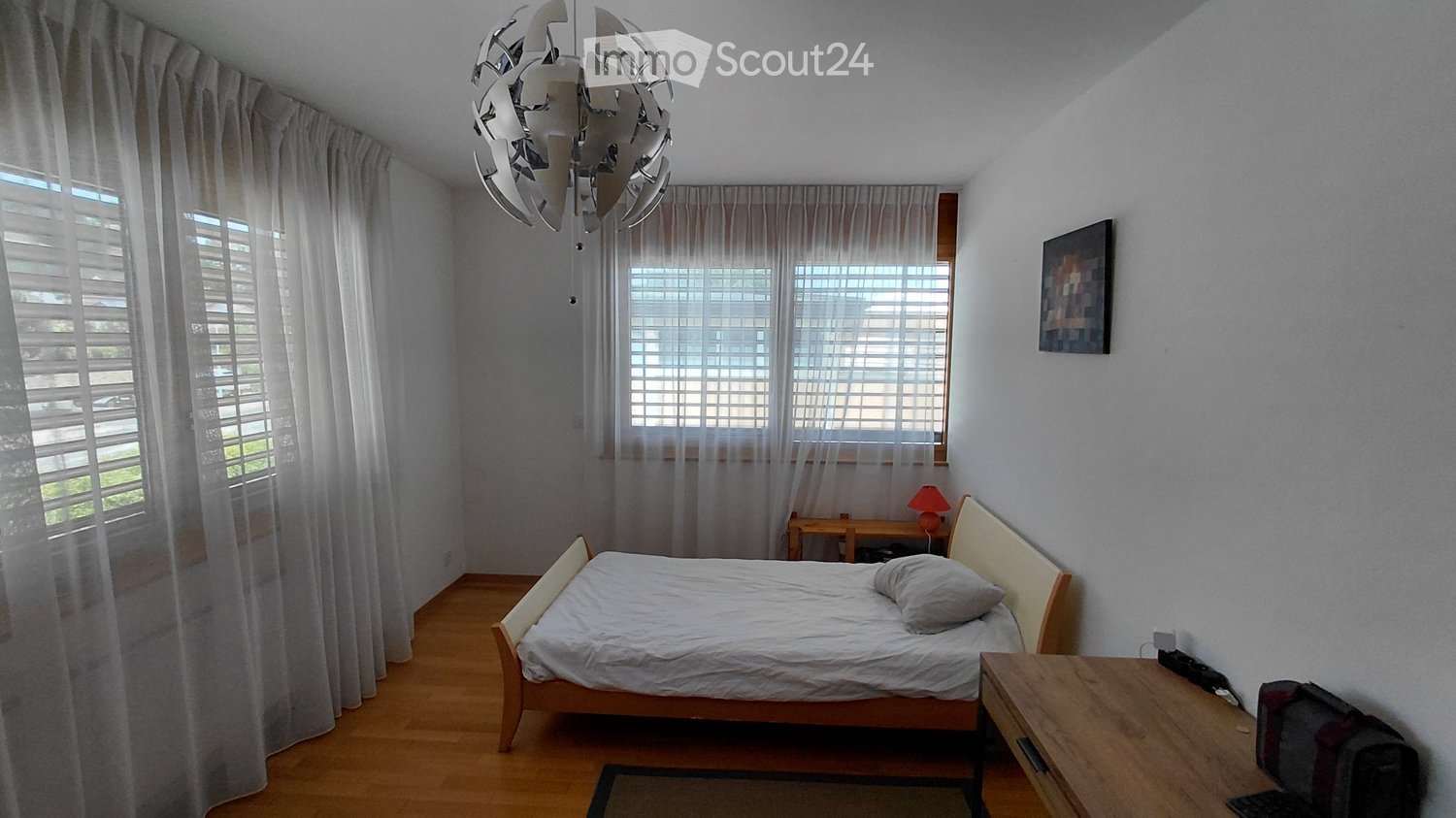 A well-lit bedroom with a wooden bed, nightstand, lamp, and a small table with a chair. The room features large windows with white curtains and a modern chandelier.