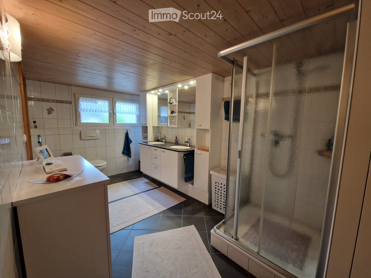 Bathroom with wooden ceiling, white cabinets, sink, mirror, shower, and tiled walls.