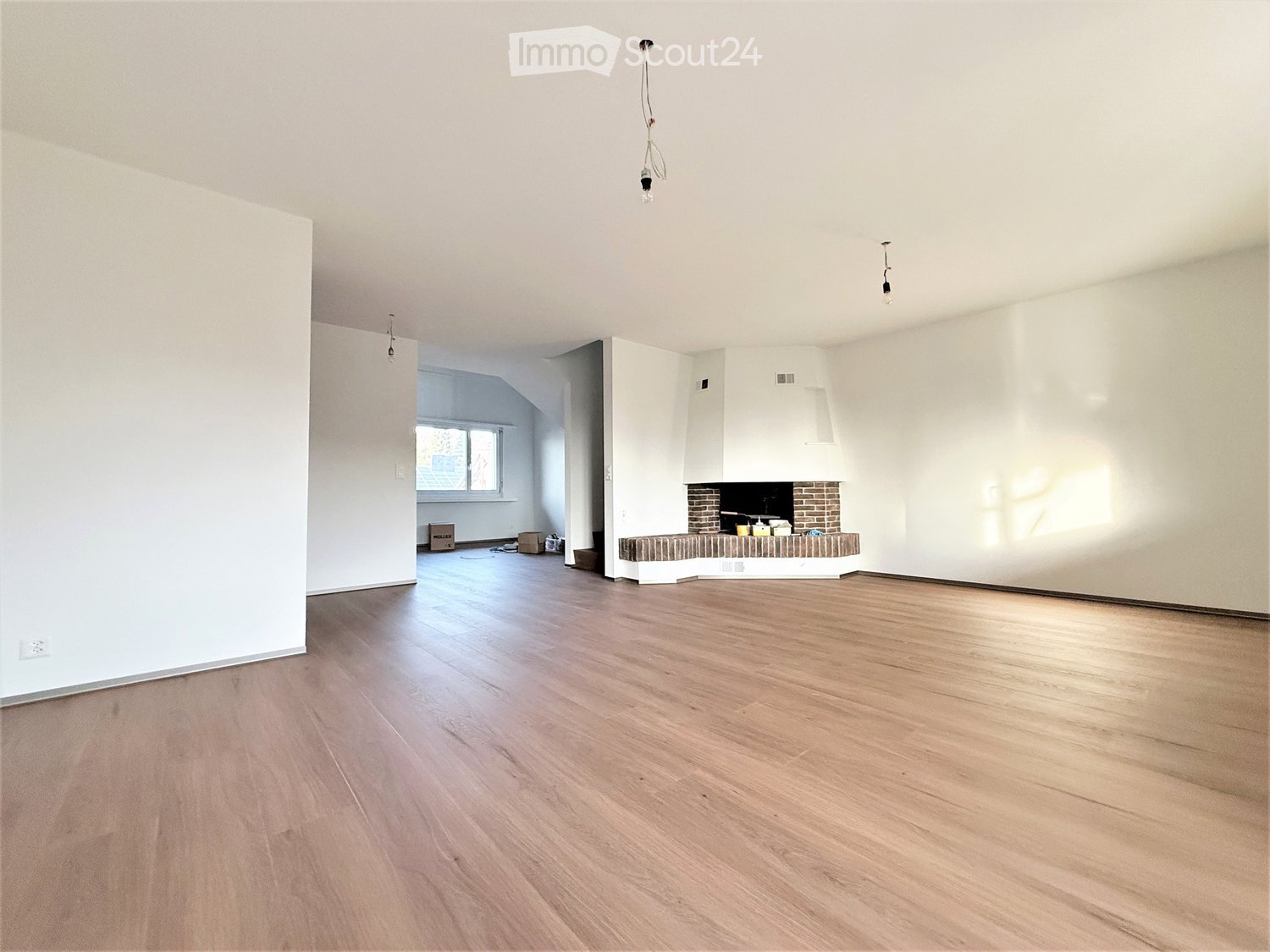 Empty room with light wooden flooring, white walls, and a fireplace with a brick surround.