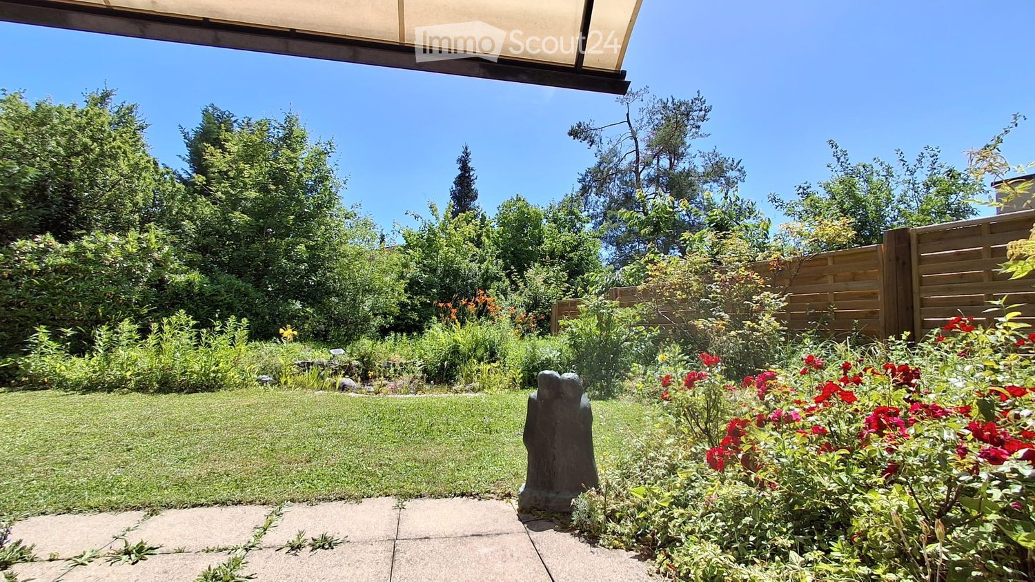 Well-maintained garden with a lawn, various flowering plants, shrubs, trees, and a wooden fence. A sculpture is placed near the pathway.