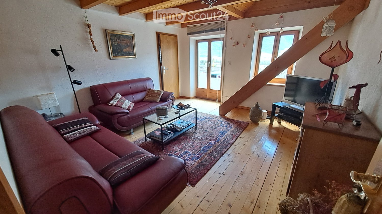 A cozy living room with wooden floors, a red leather sofa, a coffee table, a television, and a staircase leading upwards.