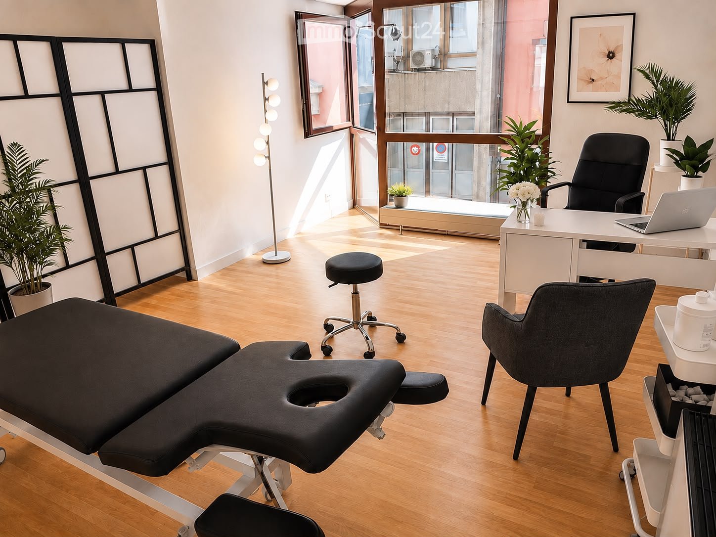 Modern, well-lit room with a therapist table, desk, laptop, chairs, and plants. Features large windows with a view and a balcony.