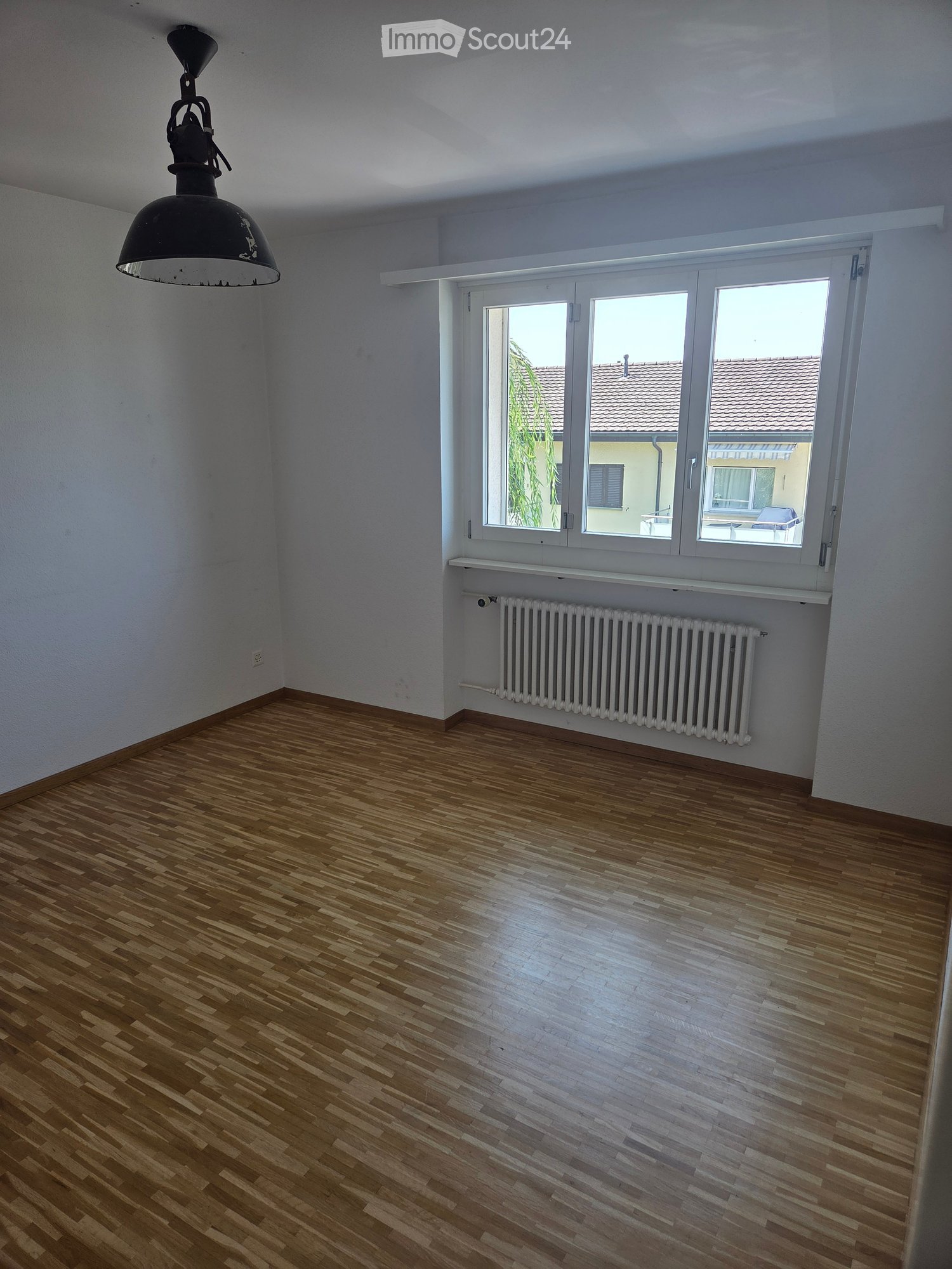 Empty room with wooden flooring, white walls, large windows, radiator, and a hanging light fixture.