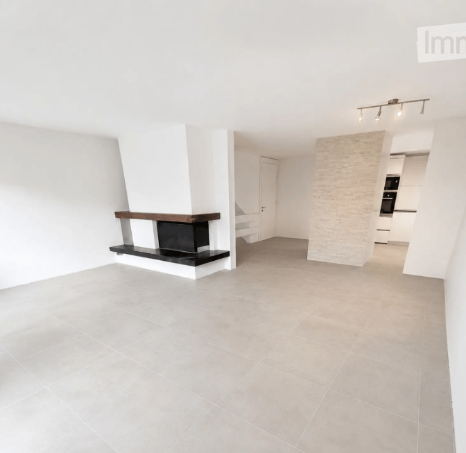 Open-plan living space with a fireplace, white walls, grey tiled floor, and recessed lighting. The room is unfurnished, showing only the fireplace and a doorway leading to another area.