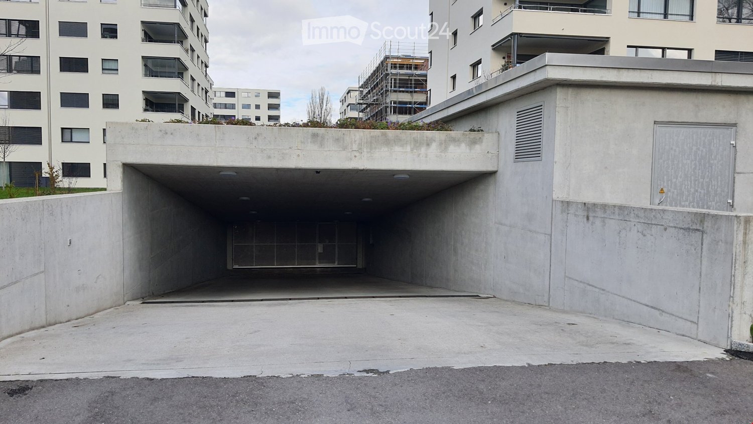 Modern concrete building entrance with a covered garage area, surrounded by other residential structures and greenery.