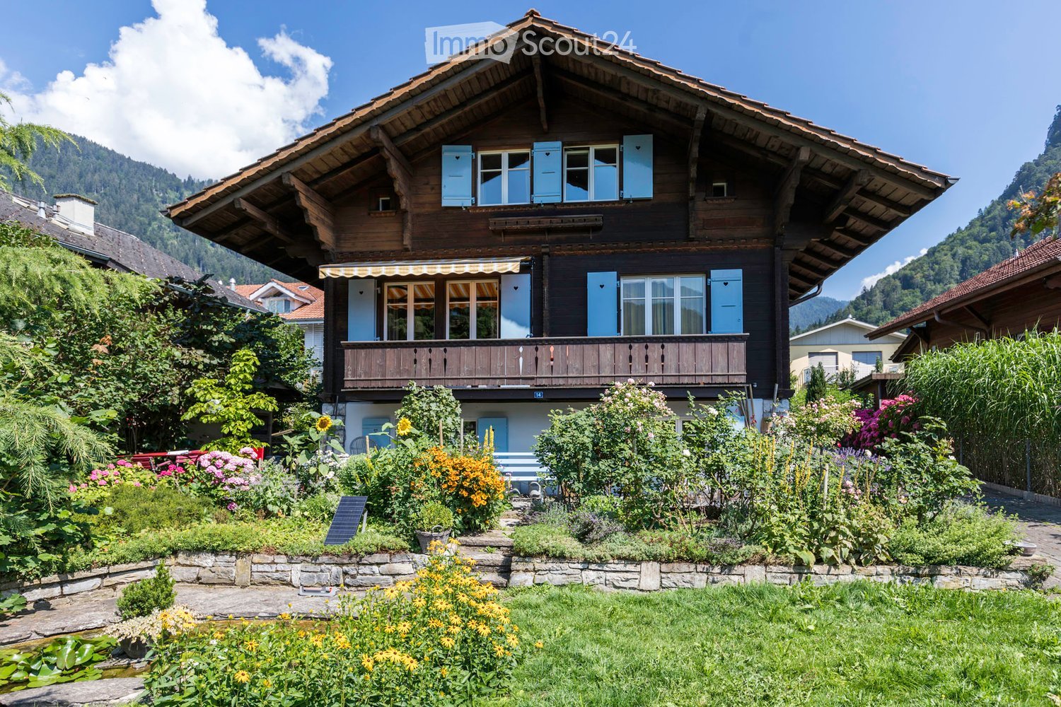 A two-story wooden house with a steep, traditional roof and light blue shutters, surrounded by a well-maintained garden with colorful flowers, shrubs, and a small pond.