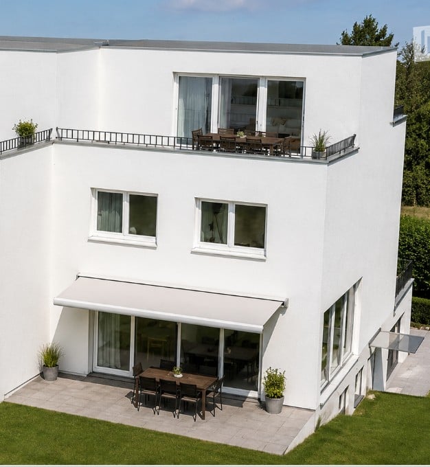 Modern two-story white house with large windows, a covered patio, and a rooftop terrace with outdoor furniture.