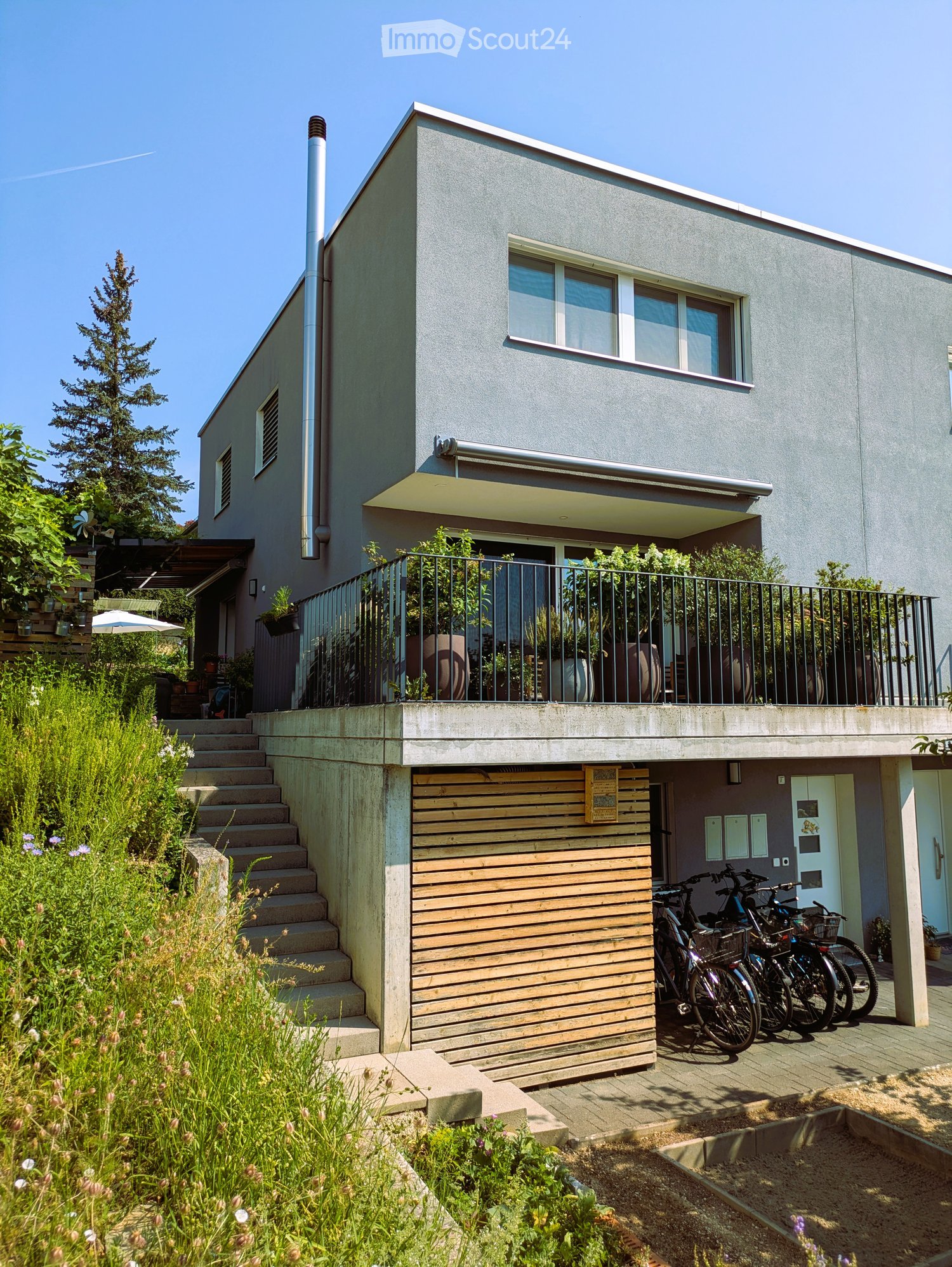 Modern two-story house with grey facade, large windows, balcony, and steps leading to a wooden garage area with bicycles parked nearby.