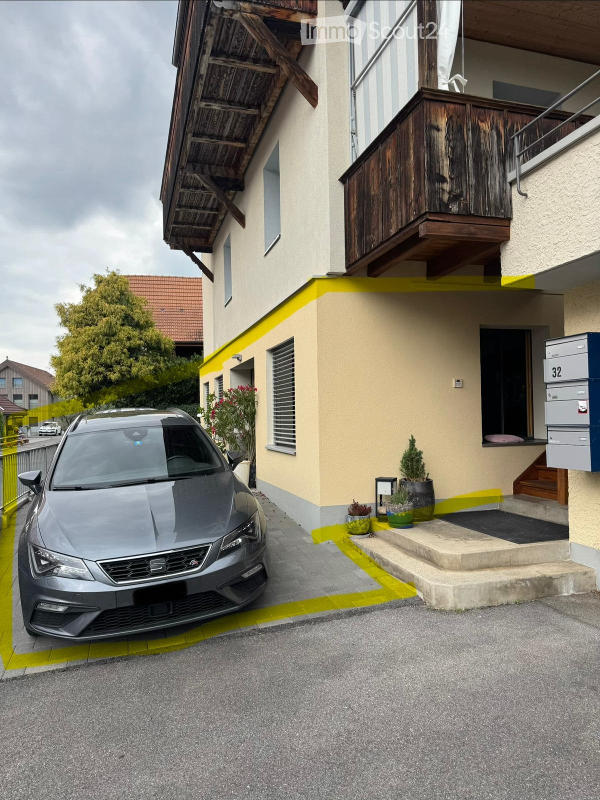 Two-story house with a balcony, grey car parked in front, small potted plants near the entrance, and a mailbox on the right side.