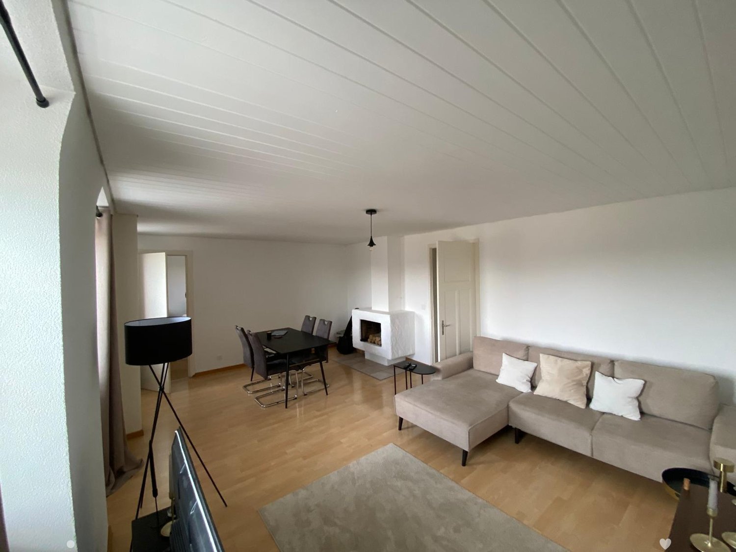Modern living room with a grey sectional sofa, coffee table, fireplace, dining area with a black table and chairs, and a television on a stand.