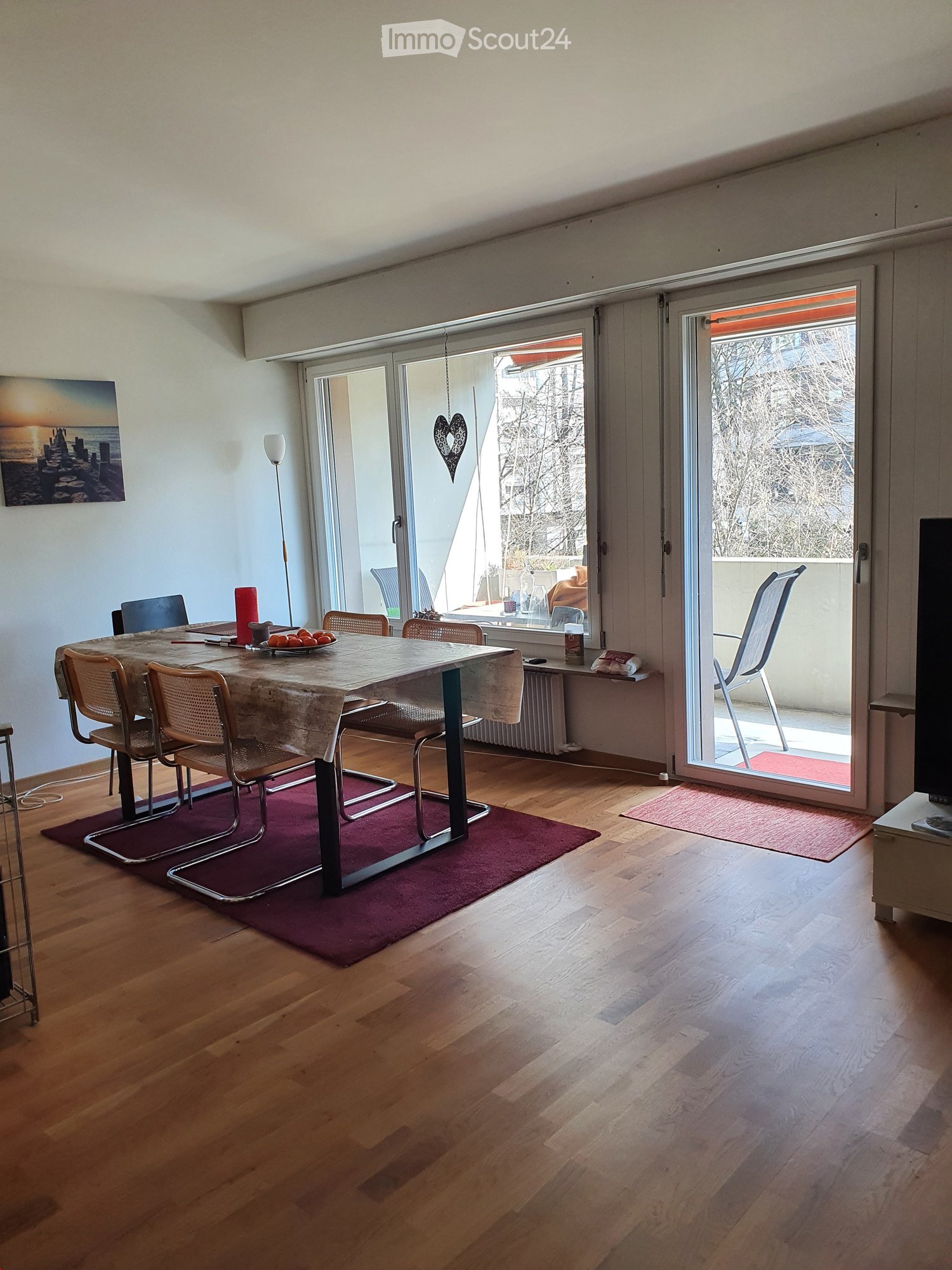 Dining room with wooden flooring, a dining table with chairs, a table runner, a floor lamp, and a television. Large sliding glass doors lead to a balcony.