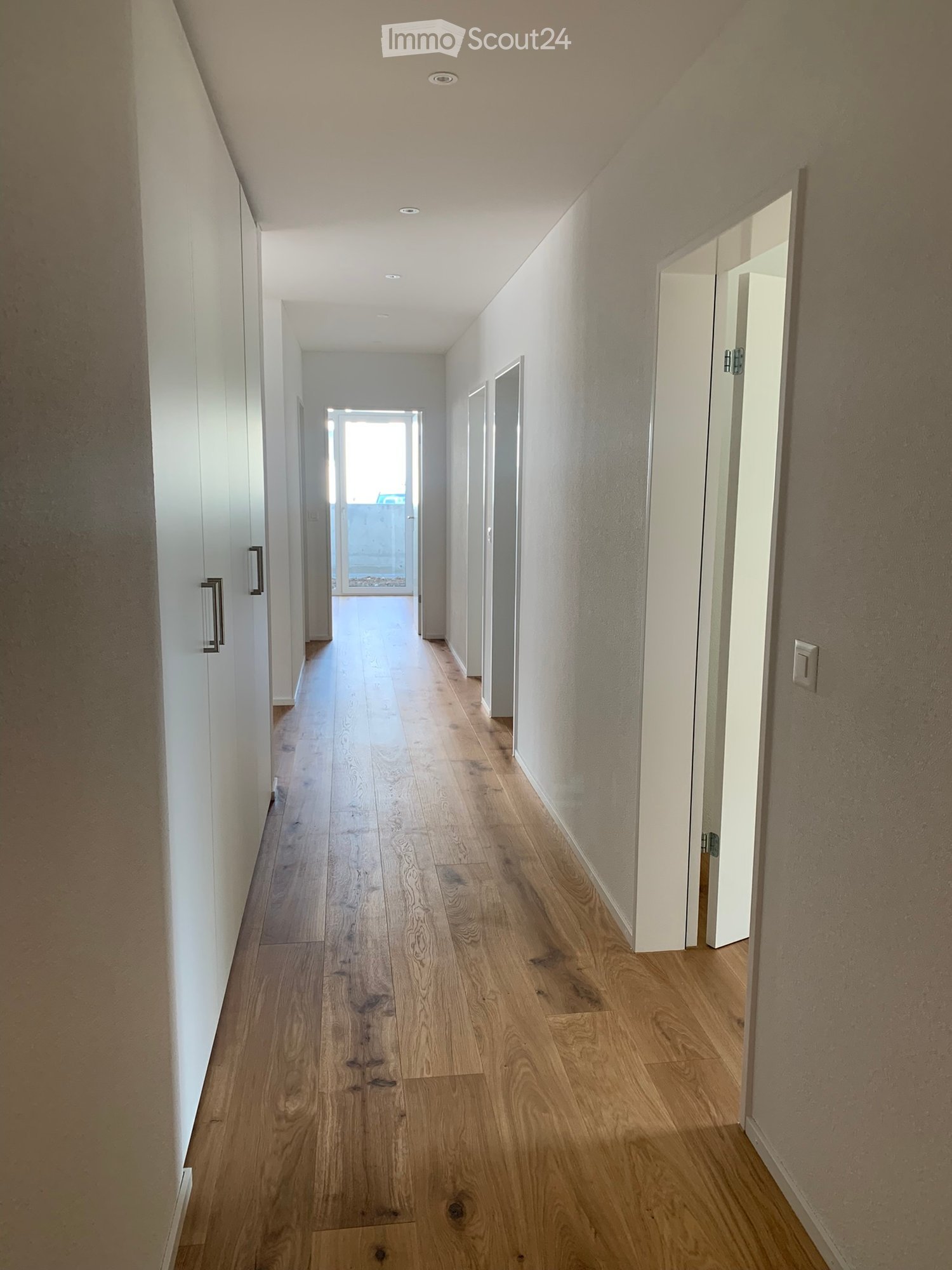 A long, narrow hallway with light wooden flooring, white walls, and recessed ceiling lighting. Doors are visible on both sides, leading to other rooms.