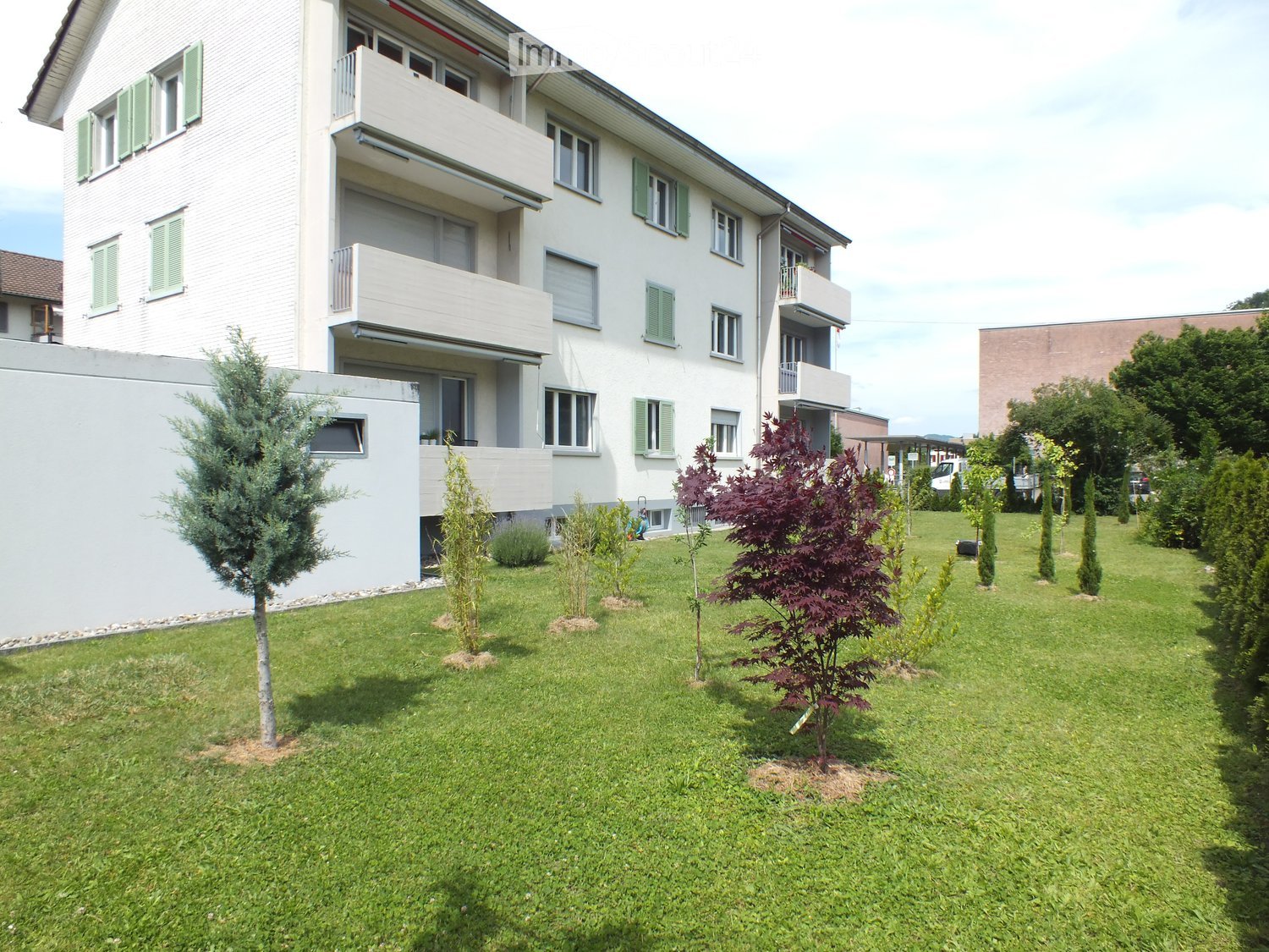 Well-maintained lawn with several small trees and shrubs, surrounded by a multi-story residential building with balconies and green shutters.
