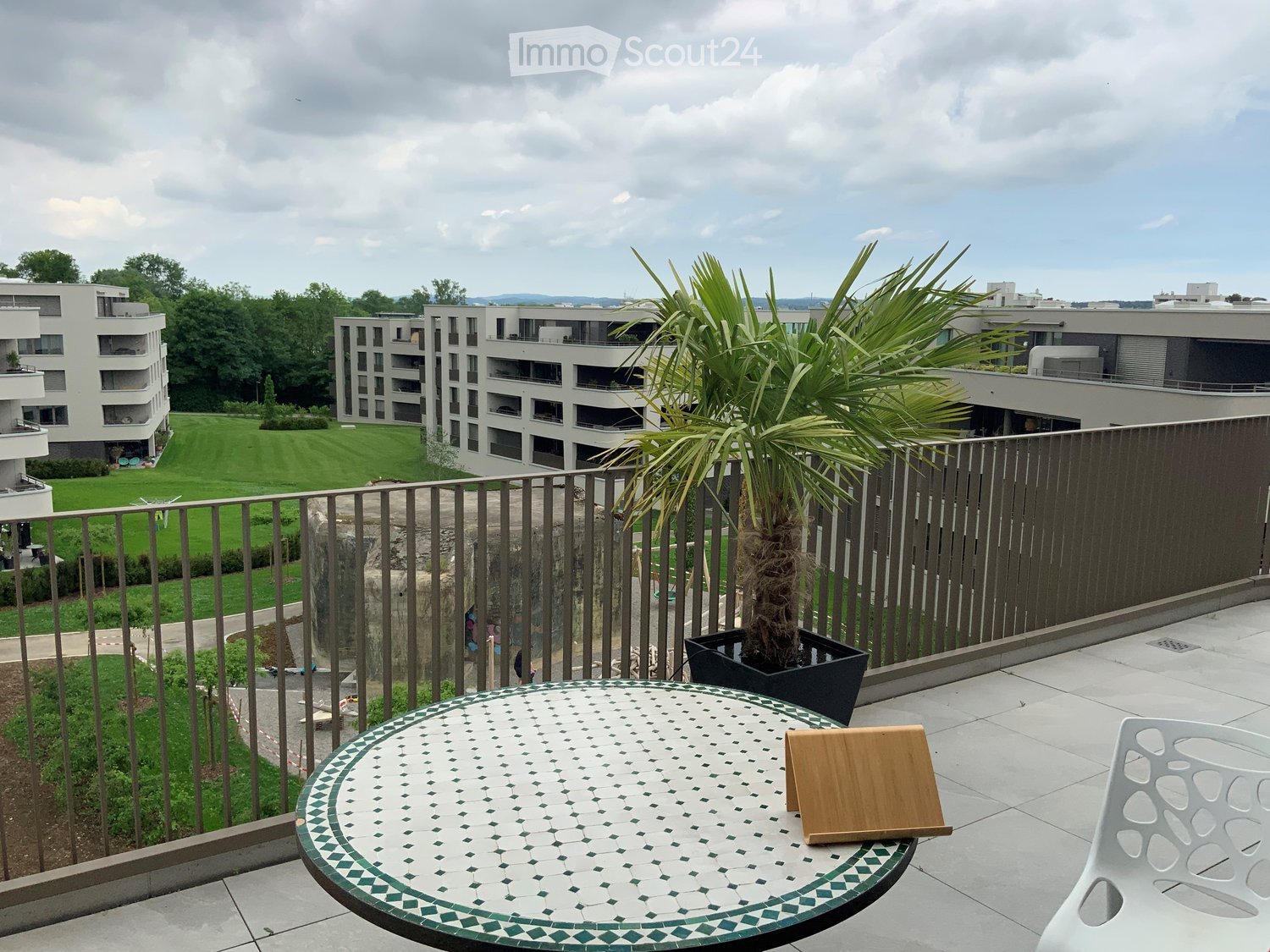 balcony with metal railings, round table with decorative design, potted palm tree, white plastic chair, background of large grass field, building with multiple balconies, cloudy sky
