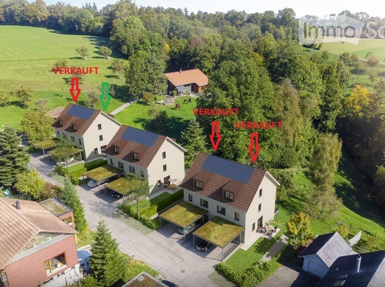 Aerial view of a residential area with multiple houses, green lawns, trees, and driveways. Some houses have solar panels on the roofs.