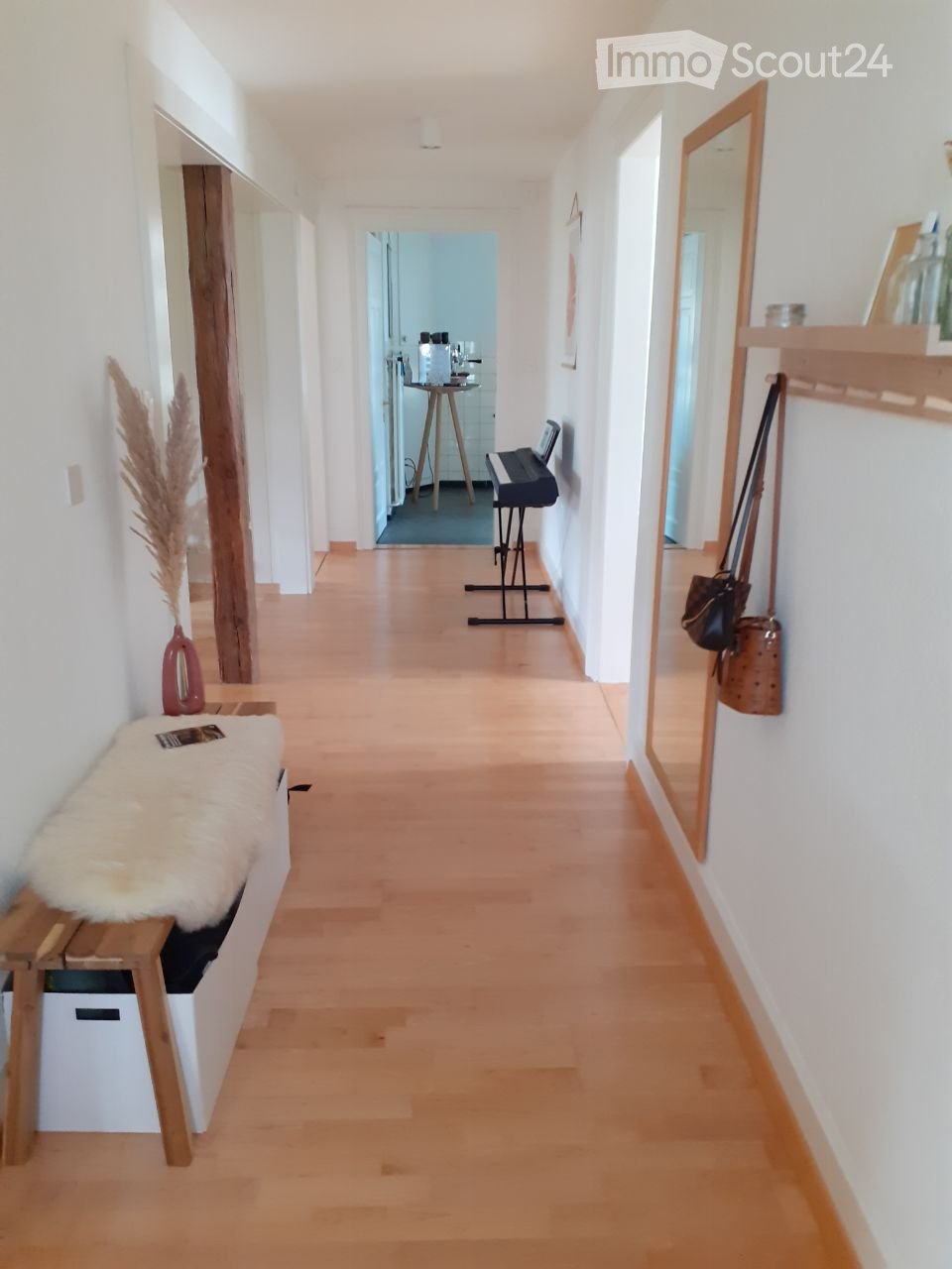 A modern hallway with light wooden flooring, white walls, and a bench with a fur throw. A small stool with glasses and a piano in the background, along with a mirror and a handbag hanging on the wall.