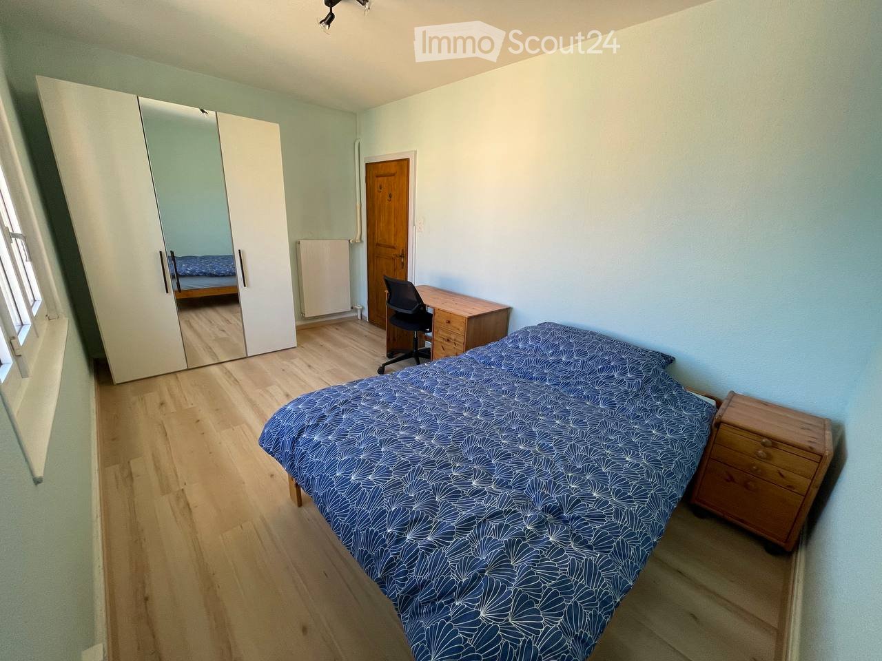 Cozy bedroom with light blue walls, wooden flooring, a single bed with blue patterned bedding, a wooden nightstand, a large mirrored wardrobe, a desk with a chair, and a radiator.