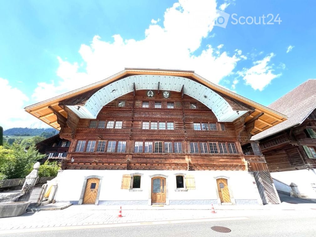 Traditional wooden chalet with a steep, multi-pitched roof and large windows, featuring a raised ground floor and multiple entrances.