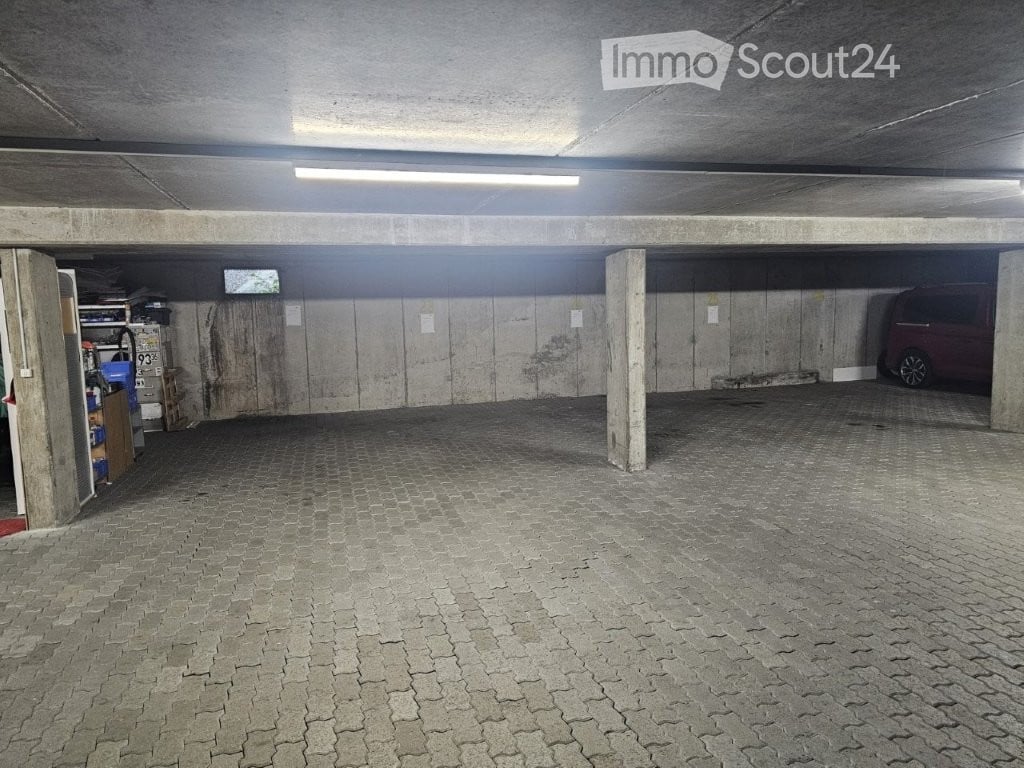Underground parking area with concrete pillars, tiled floor, and storage shelves on the left side. A parked van is visible on the right.