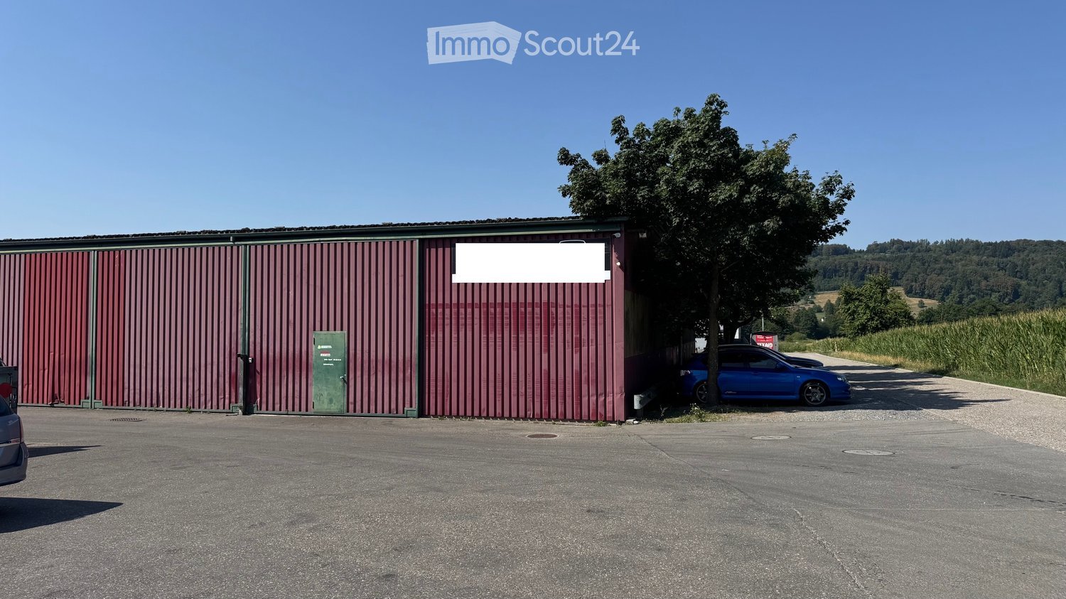 A large industrial-style building with red corrugated metal walls and a green door, situated in a rural area with a tree and a blue car parked nearby.