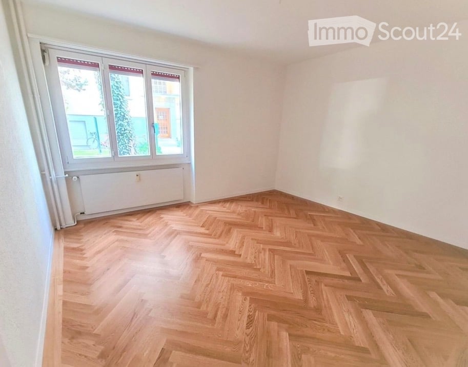 Empty room with herringbone wooden flooring, white walls, large window with white frame and red blinds, radiator below window.